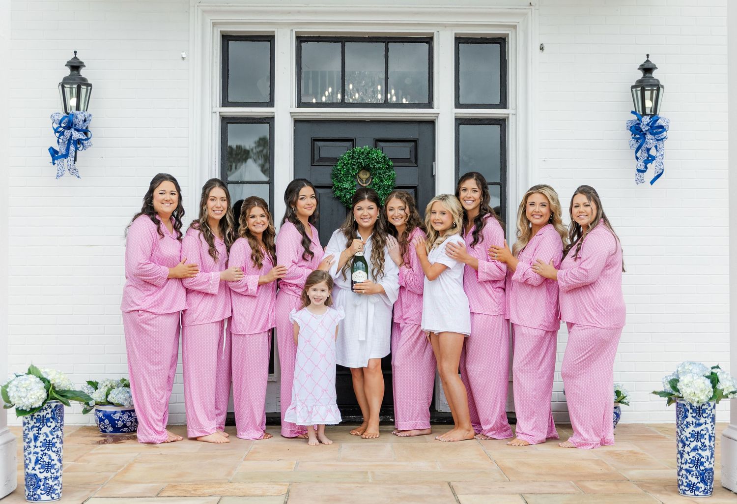 Bridal party in pink pajamas posing with bride in white robe outside white venue with blue bows.