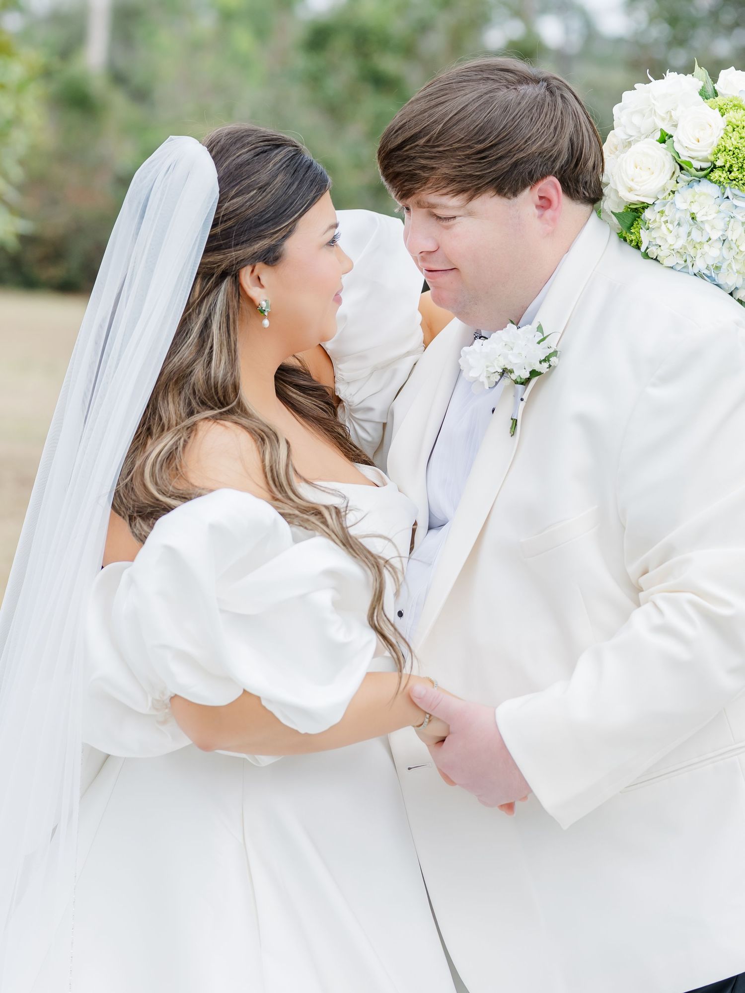 Bride and groom in white wedding attire share romantic moment gazing at each other outdoors.