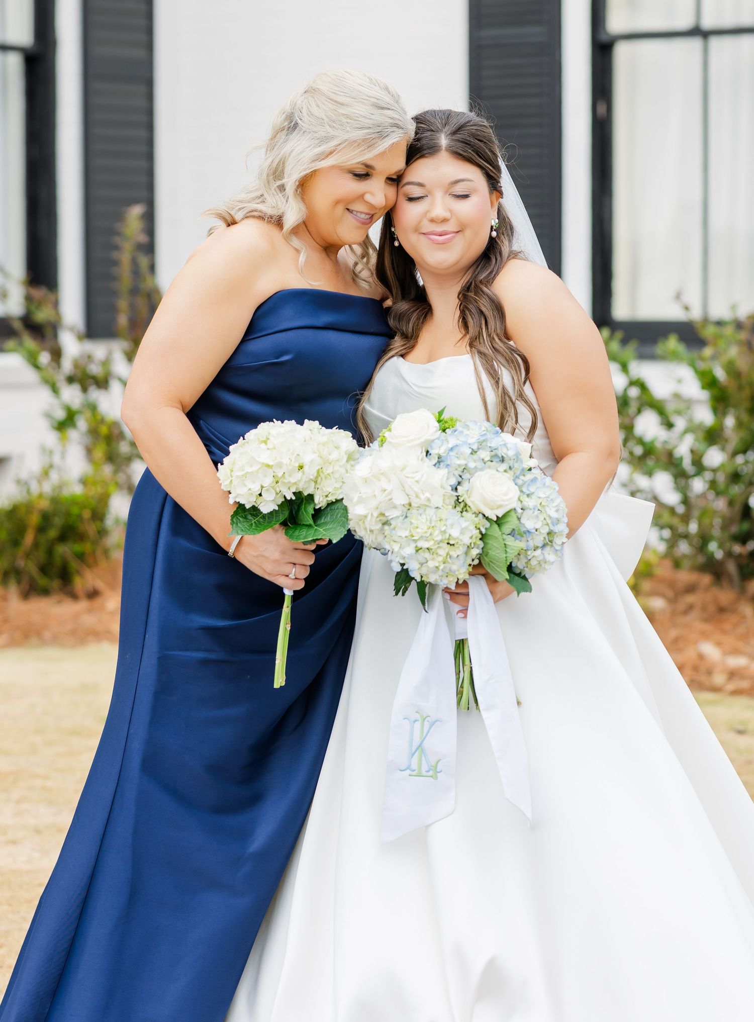 Bride in white gown and bridesmaid in navy blue dress holding white hydrangea bouquets at outdoor wedding.
