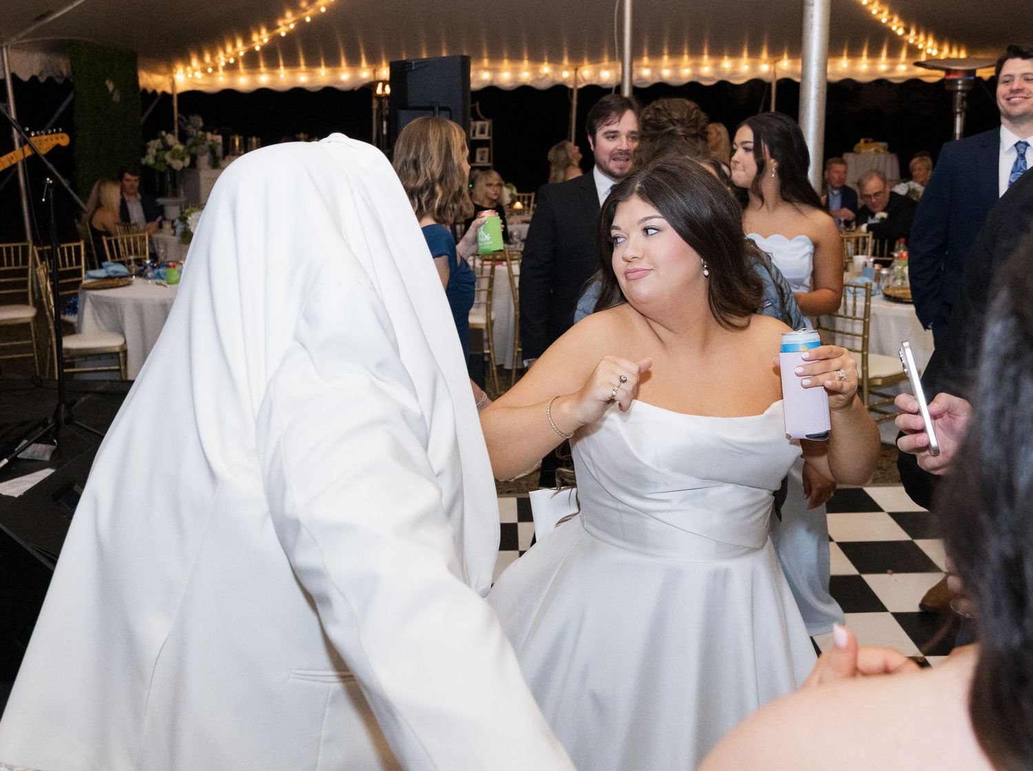 Bride in off-shoulder gray gown dancing with guest in white suit at elegant tented reception.