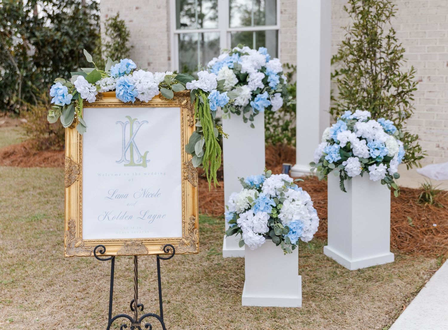 Outdoor wedding welcome sign with blue hydrangea floral arrangements on white pedestals in a garden setting.