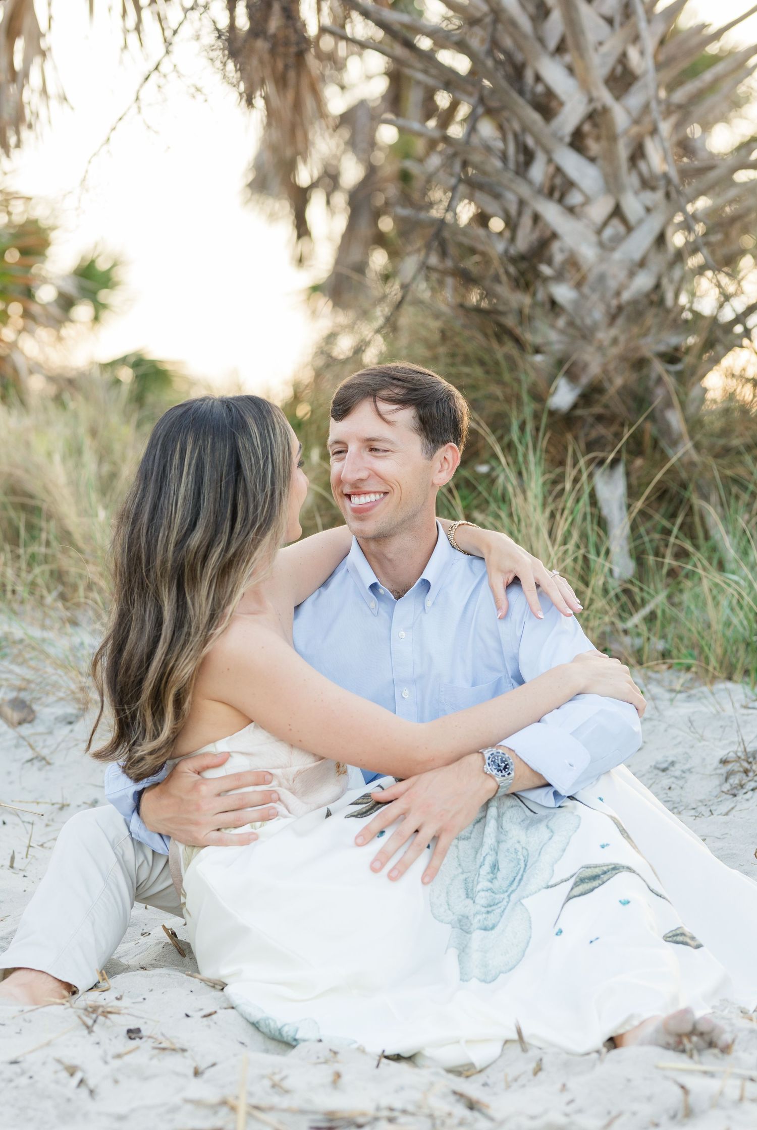 Couple sitting on beach sand embracing and smiling at each other with palm trees in background.