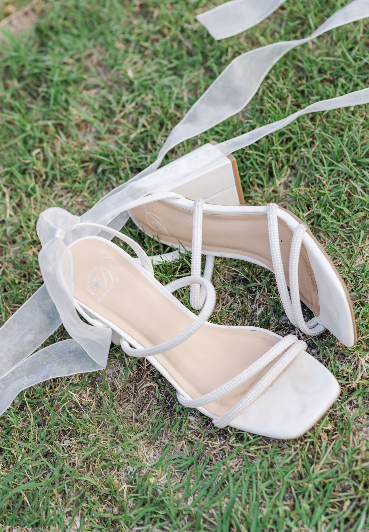 White strappy bridal heels with ribbon laces resting on green grass for a wedding day detail shot.