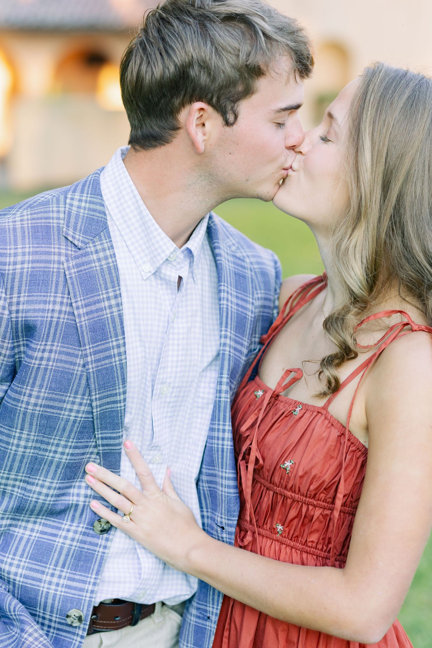 A romantic couple shares a tender kiss outdoors, with the woman wearing a red dress and man in a blue plaid shirt.