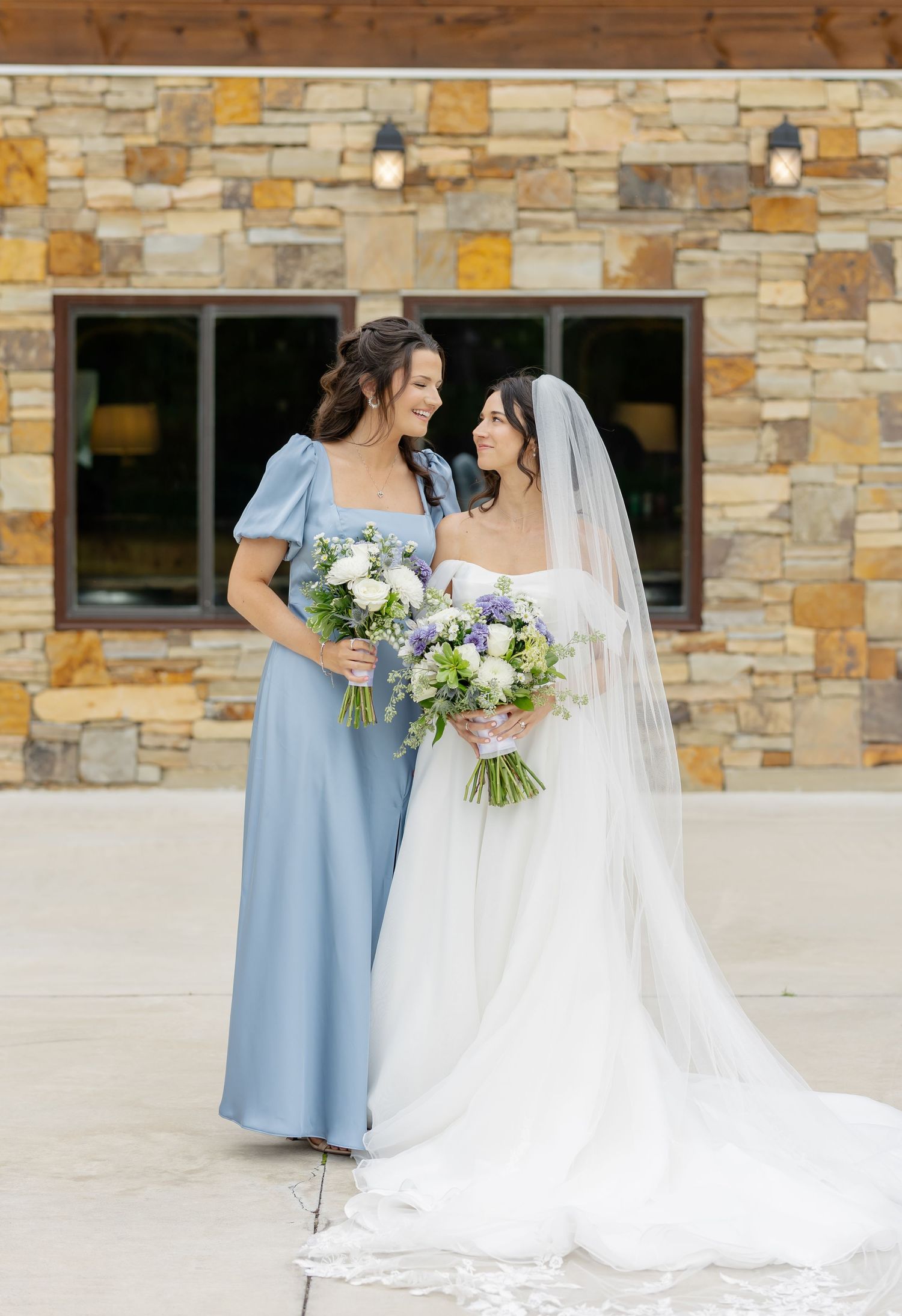 Wedding party members in light blue dresses and white gown pose against stone wall backdrop.