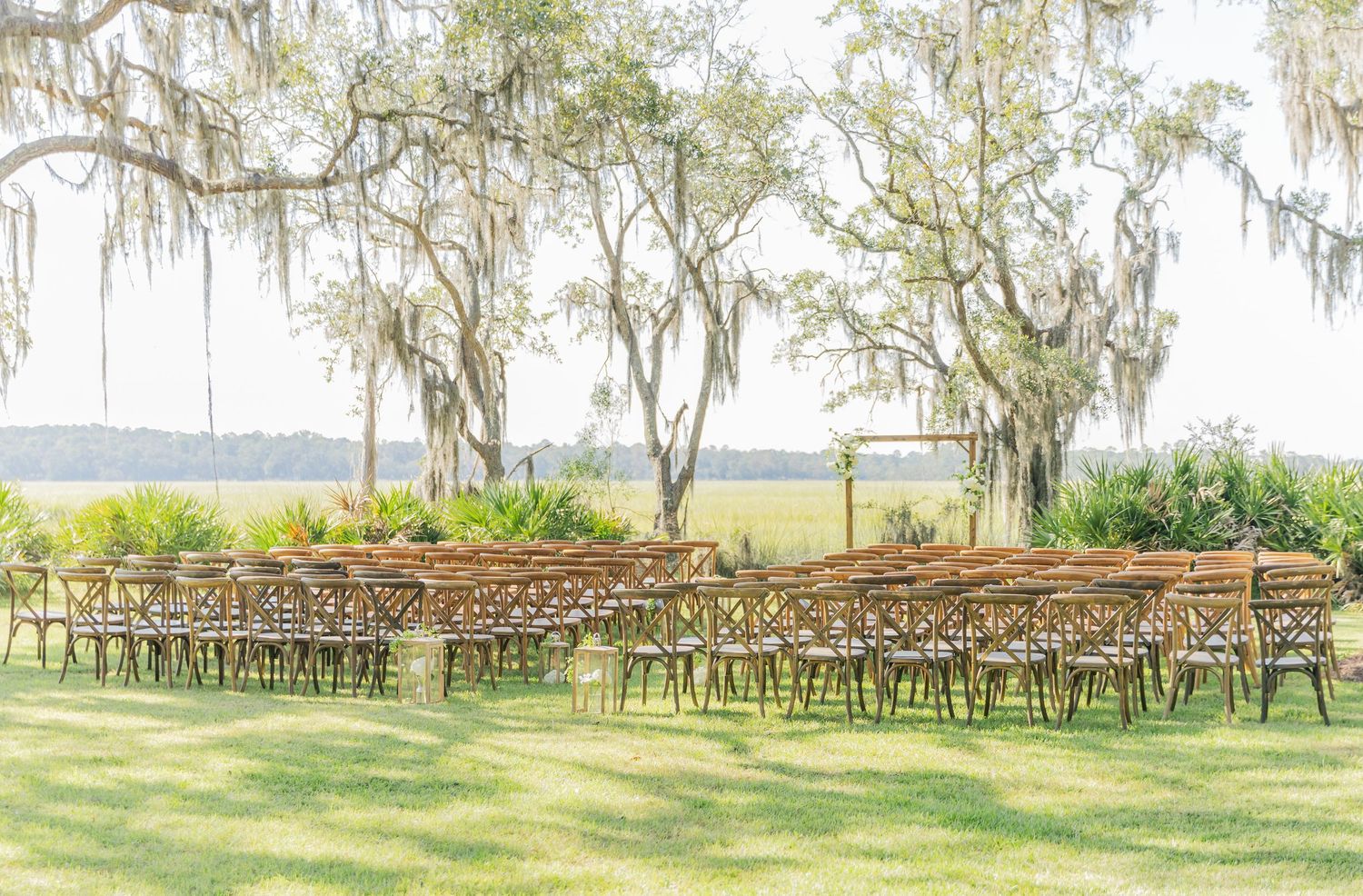 Wedding ceremony setup with wooden chairs on a lawn surrounded by moss-covered oak trees and marsh views.