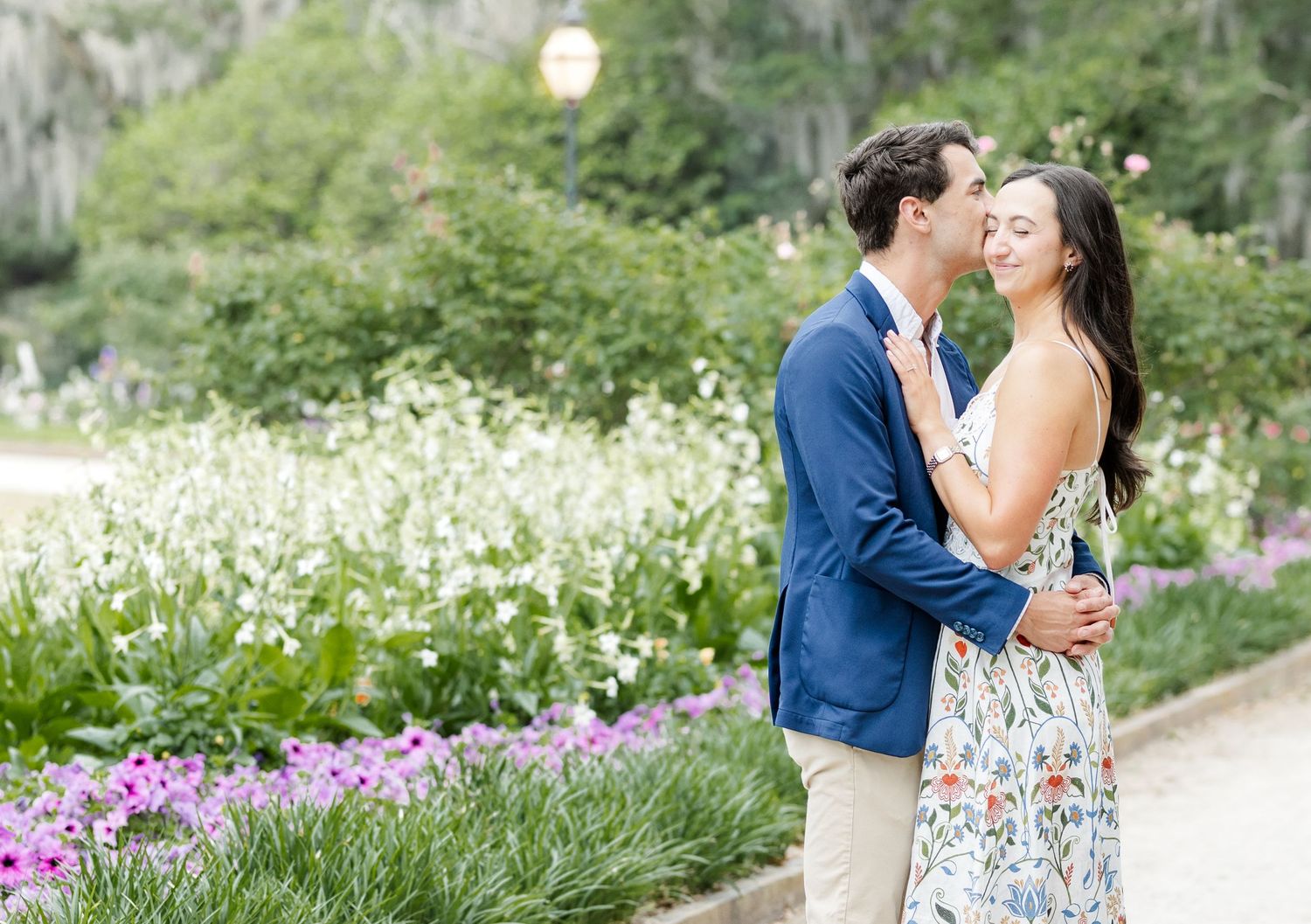 Couple embracing and kissing in a lush garden with colorful flowers, man in blue blazer, woman in floral dress.