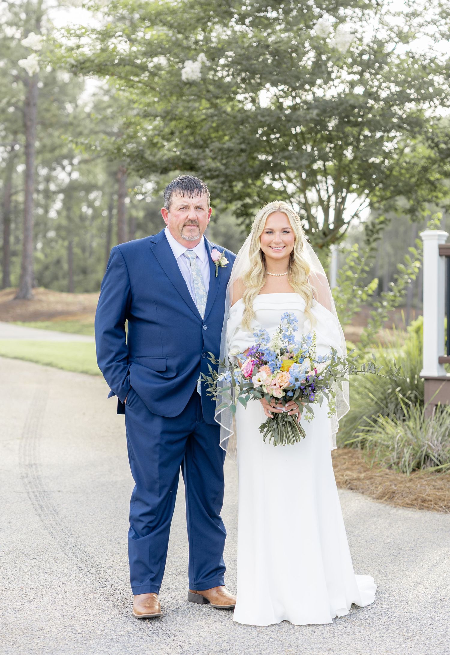 Bride with colorful bouquet poses with man in navy suit on tree-lined path.