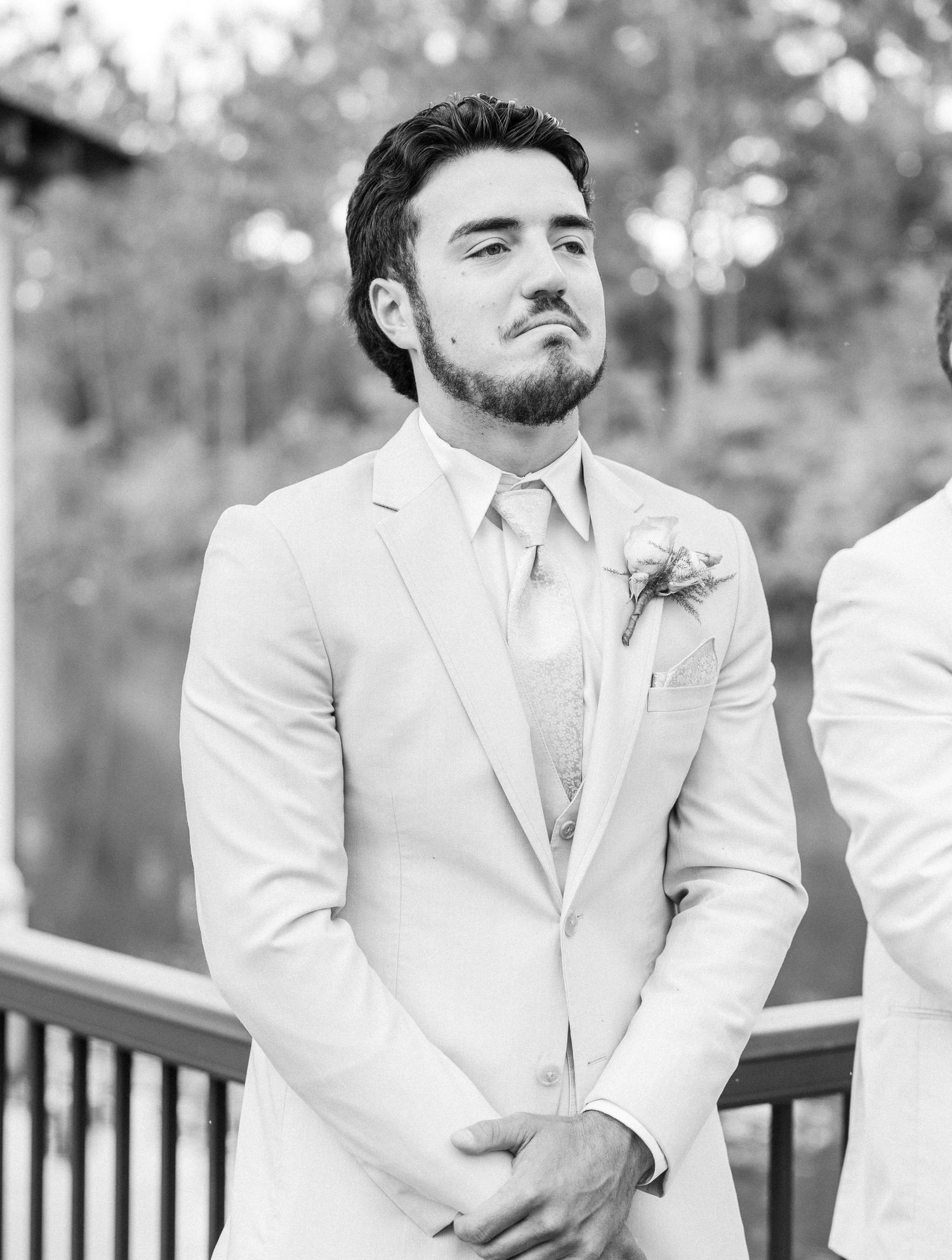 An emotional groom in a white suit watches his bride walk down the aisle in black and white.