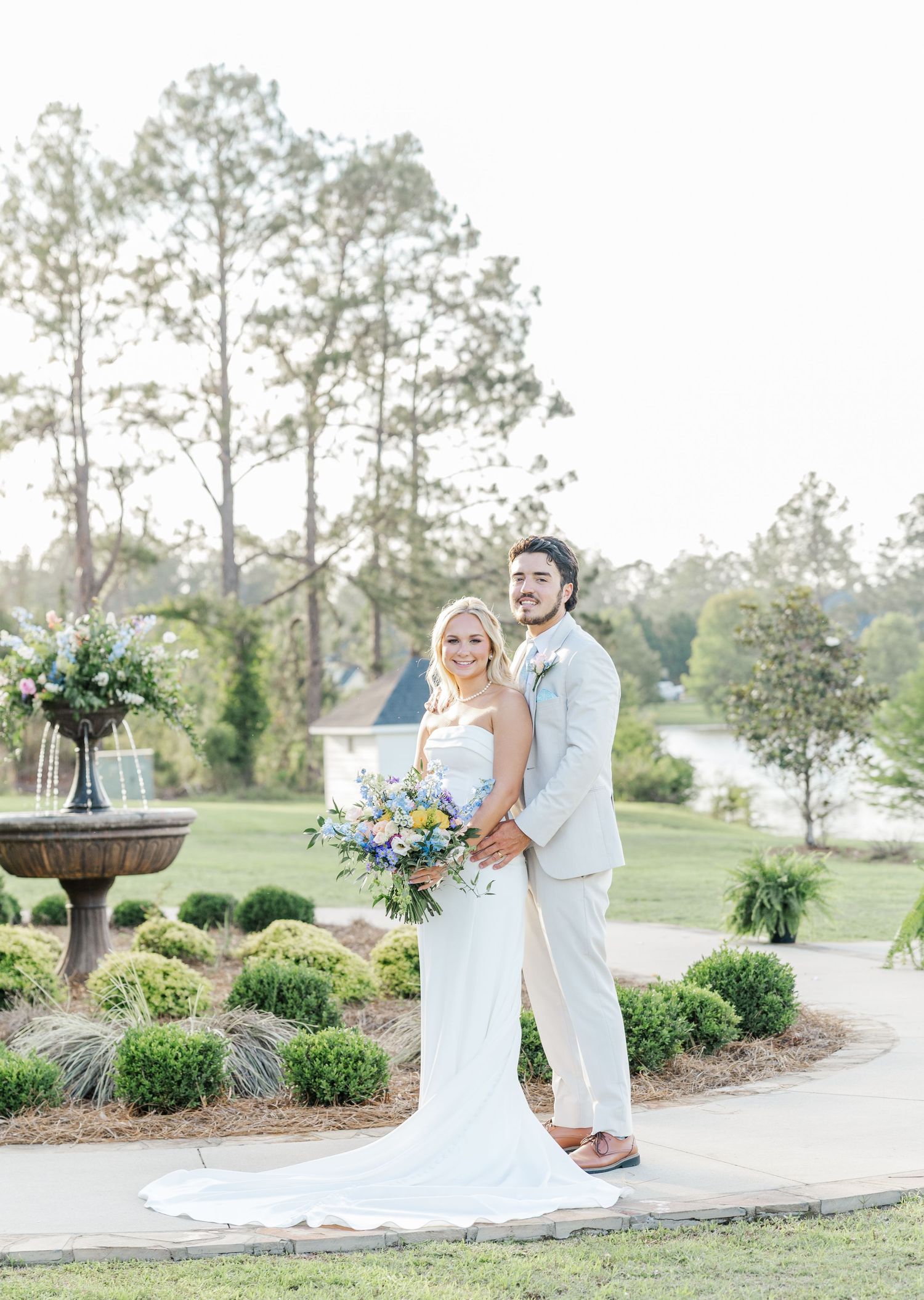 Bride and groom pose together on a garden path holding a colorful bouquet at their outdoor wedding.