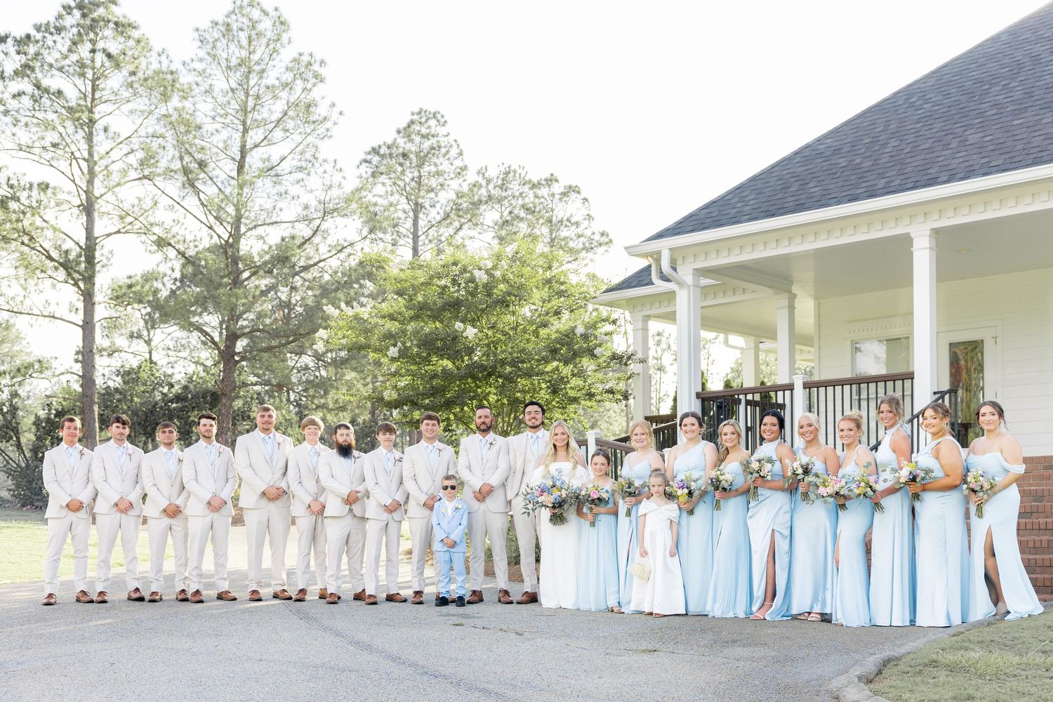 Large wedding party in light blue and beige attire posing outside white venue building.
