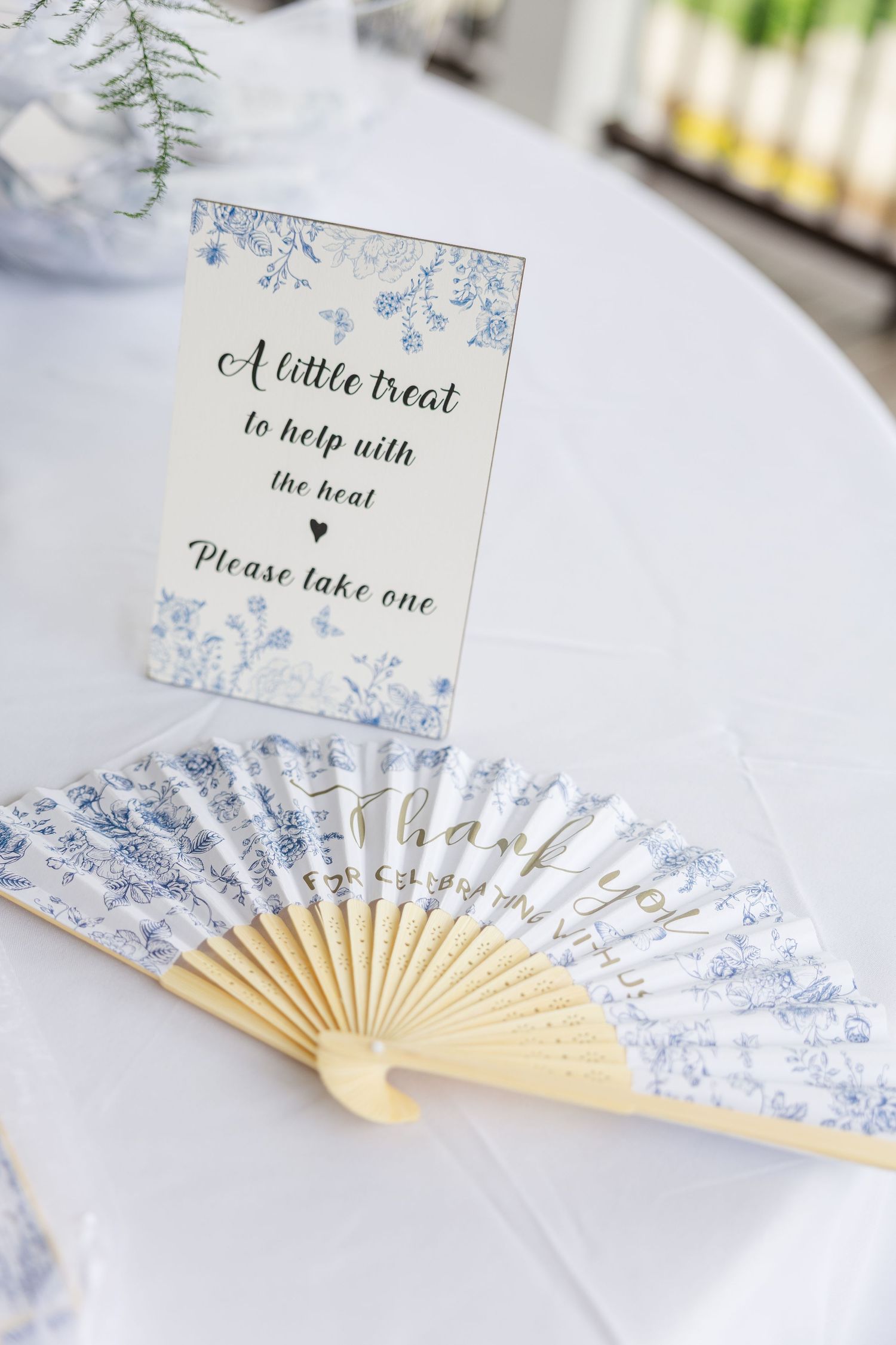 Blue and white floral paper hand fans displayed on a table with a sign reading A little treat to help with the heat please take one.