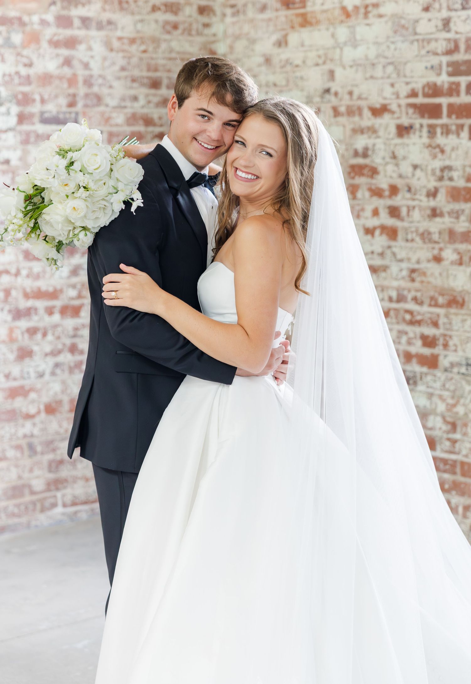 Smiling bride and groom pose together in front of brick wall, bride holding white bouquet in elegant wedding attire.