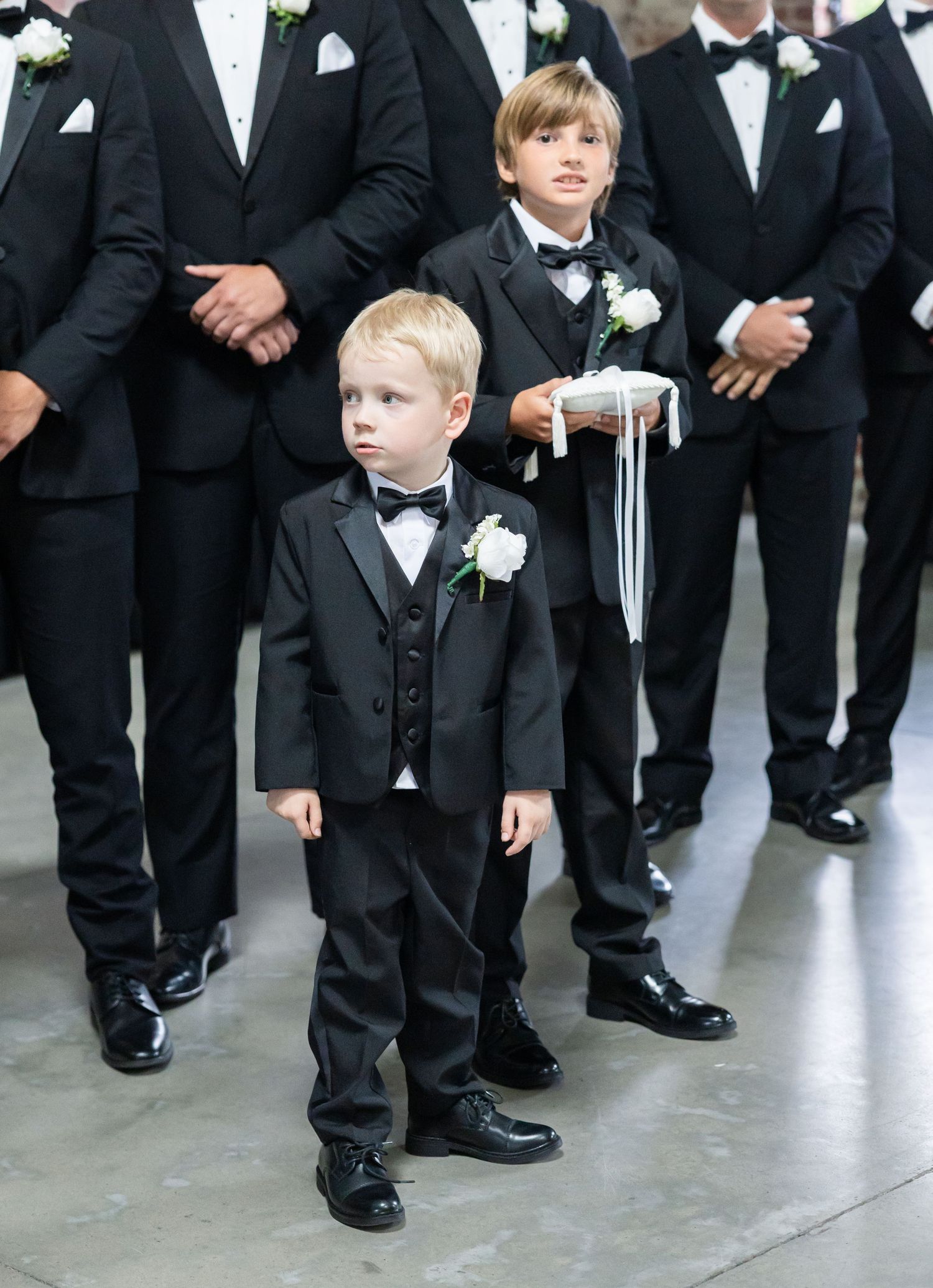Two young ring bearers in black tuxedos standing with groomsmen at wedding ceremony.