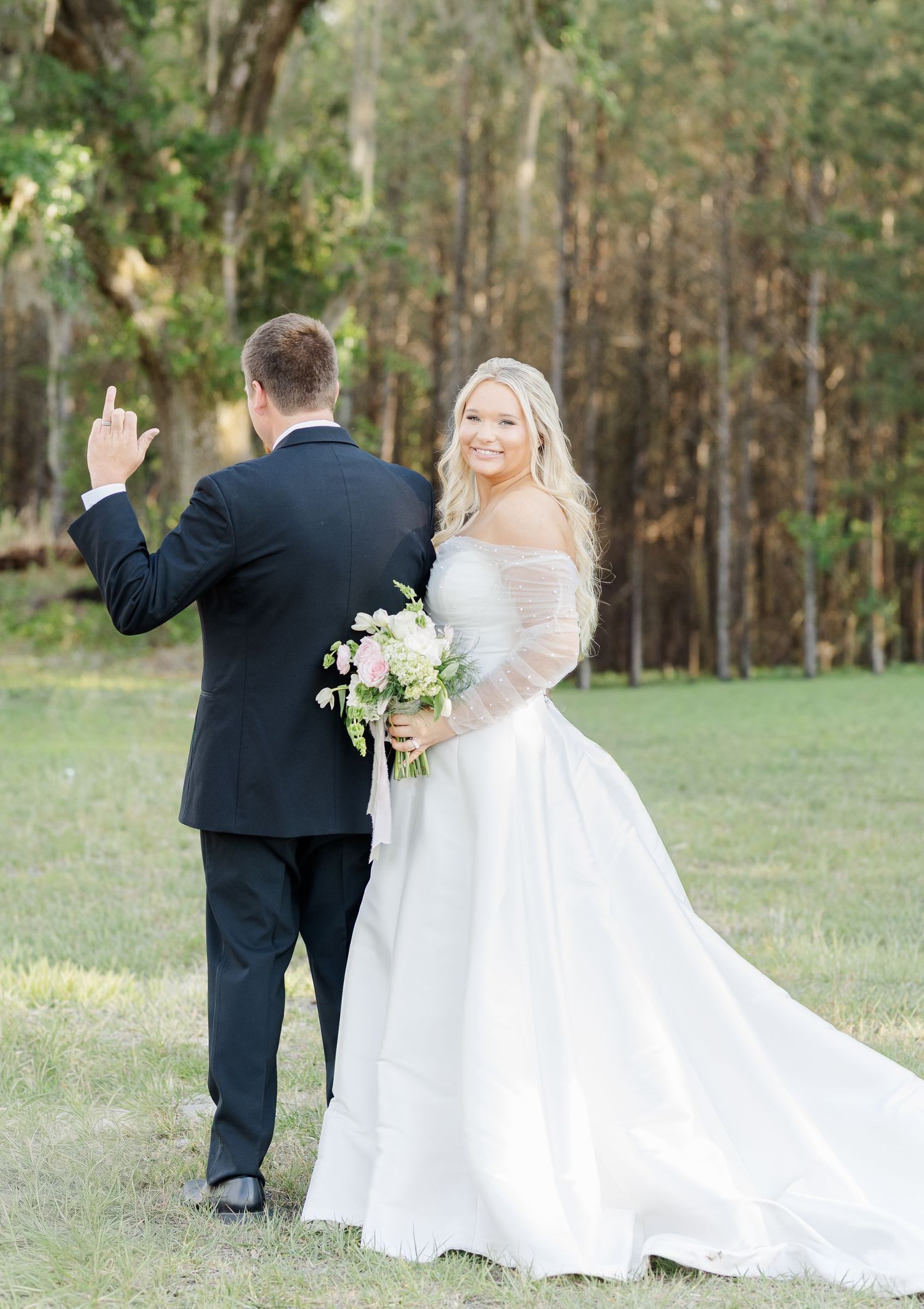 Smiling bride in white ball gown holds bouquet as groom waves goodbye in outdoor forest setting.