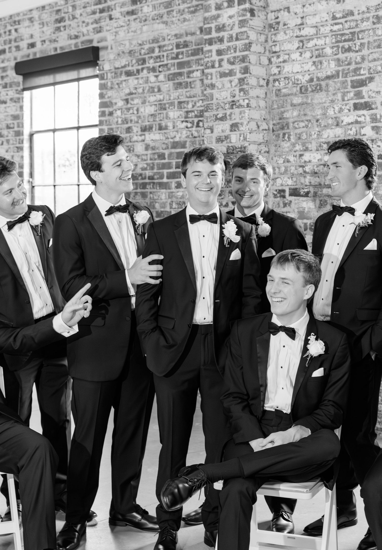 Black and white photo of groom laughing with groomsmen in tuxedos against brick wall backdrop.