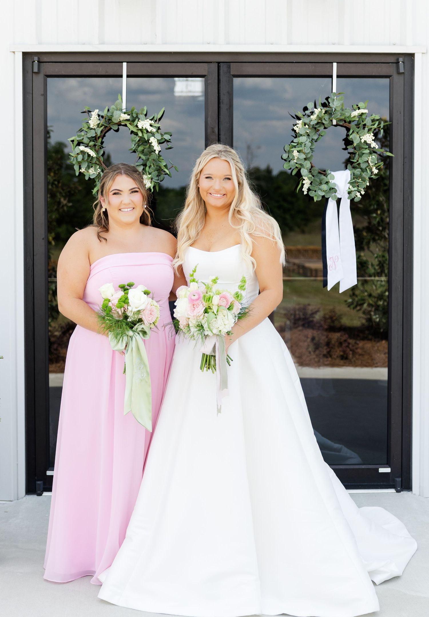 Bride in white gown and bridesmaid in pink dress holding bouquets before floral wreath doors.