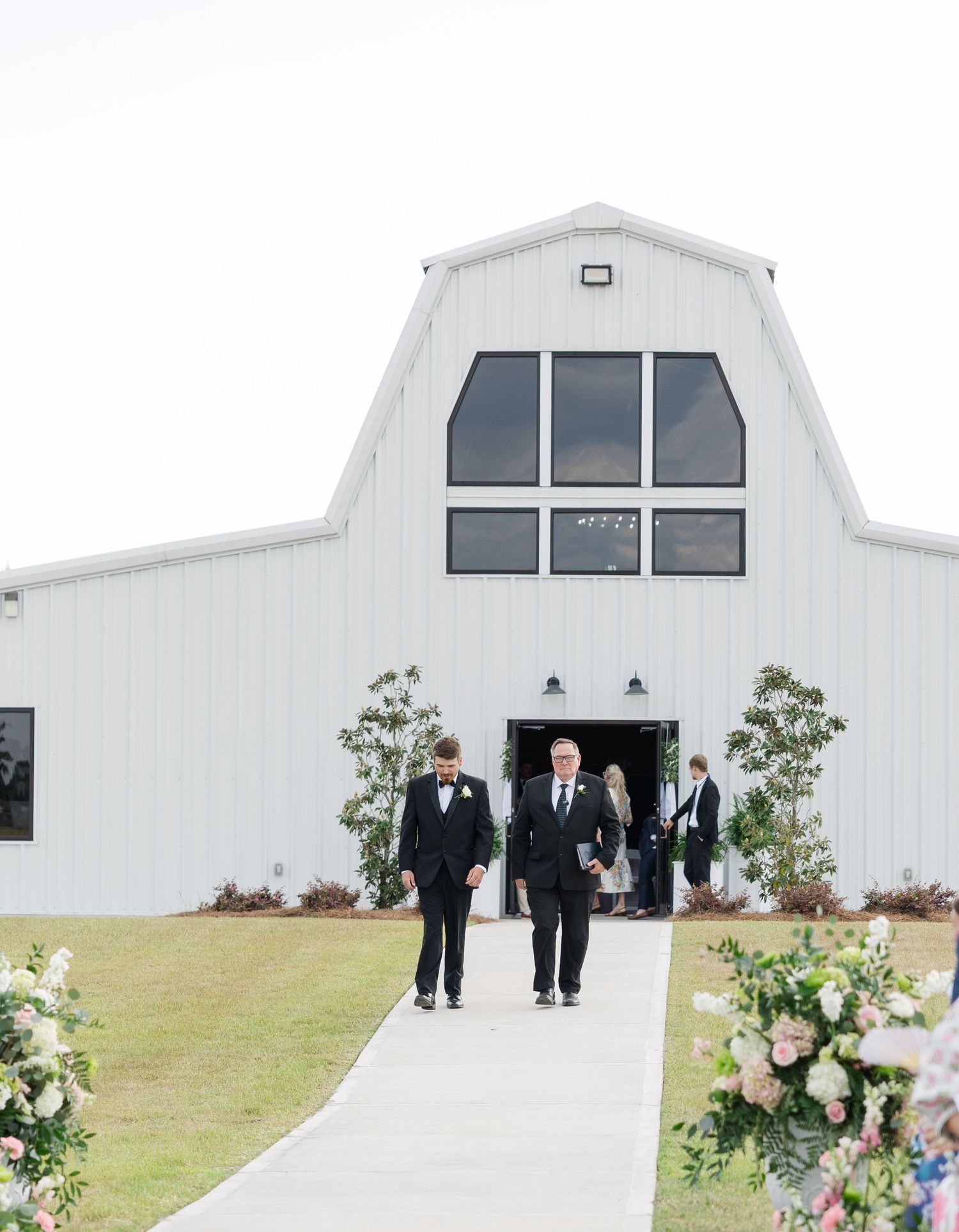 Two groomsmen in black tuxedos walking down the outdoor ceremony aisle from a white barn wedding venue.