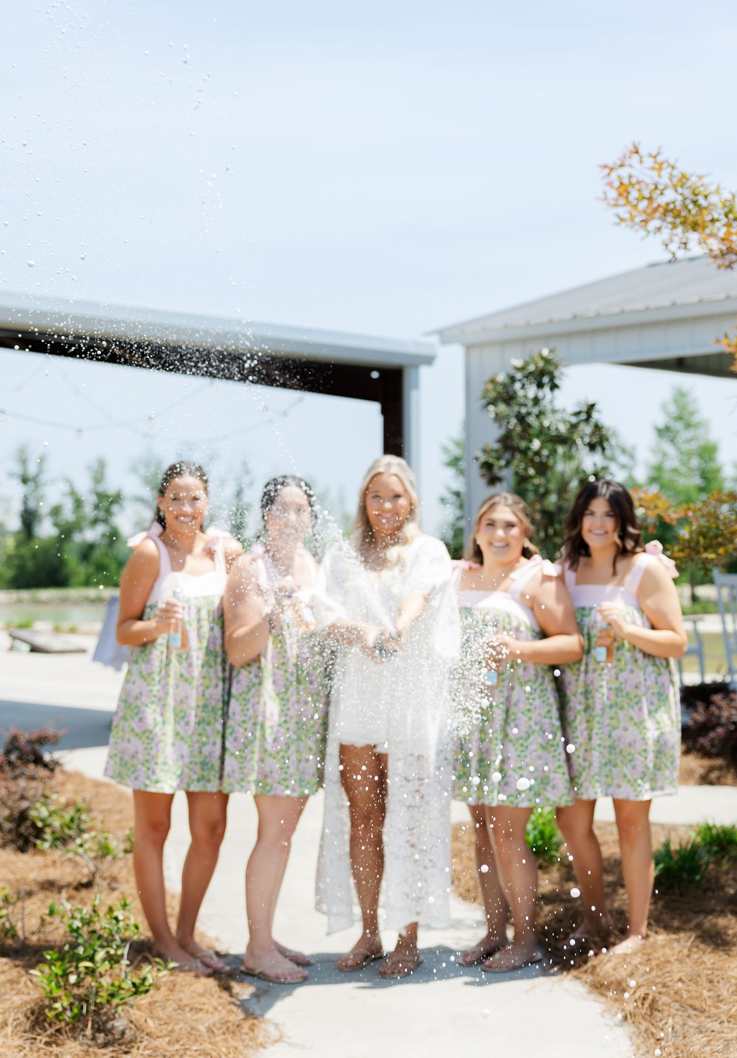 Bride in lace gown celebrates with four bridesmaids in floral dresses as confetti flies outdoors.