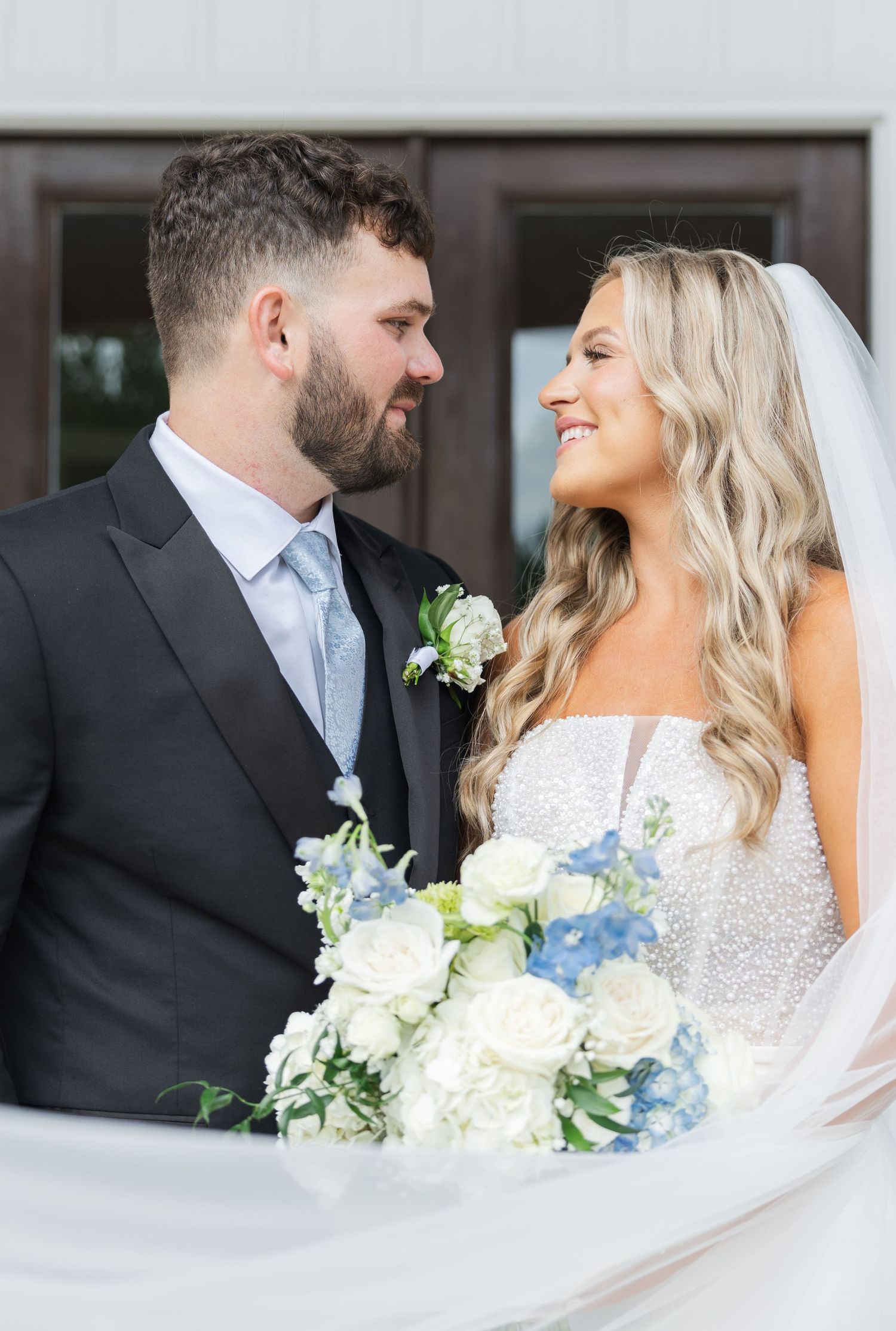Bride and groom gazing at each other, bride holding white and blue bouquet, groom in black suit with light blue tie.