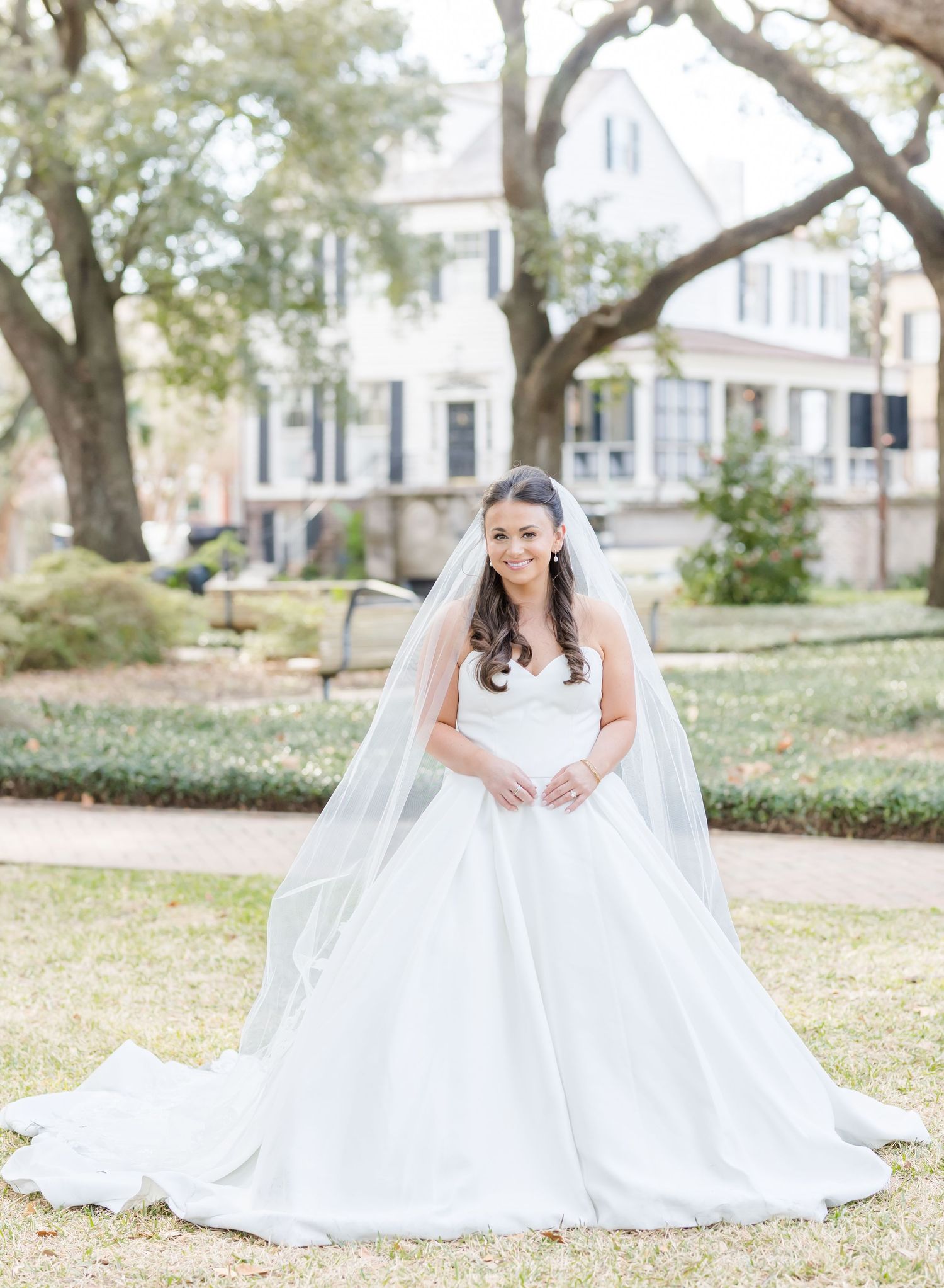 A bride in a flowing white ballgown poses in a garden setting with historic buildings in the background.