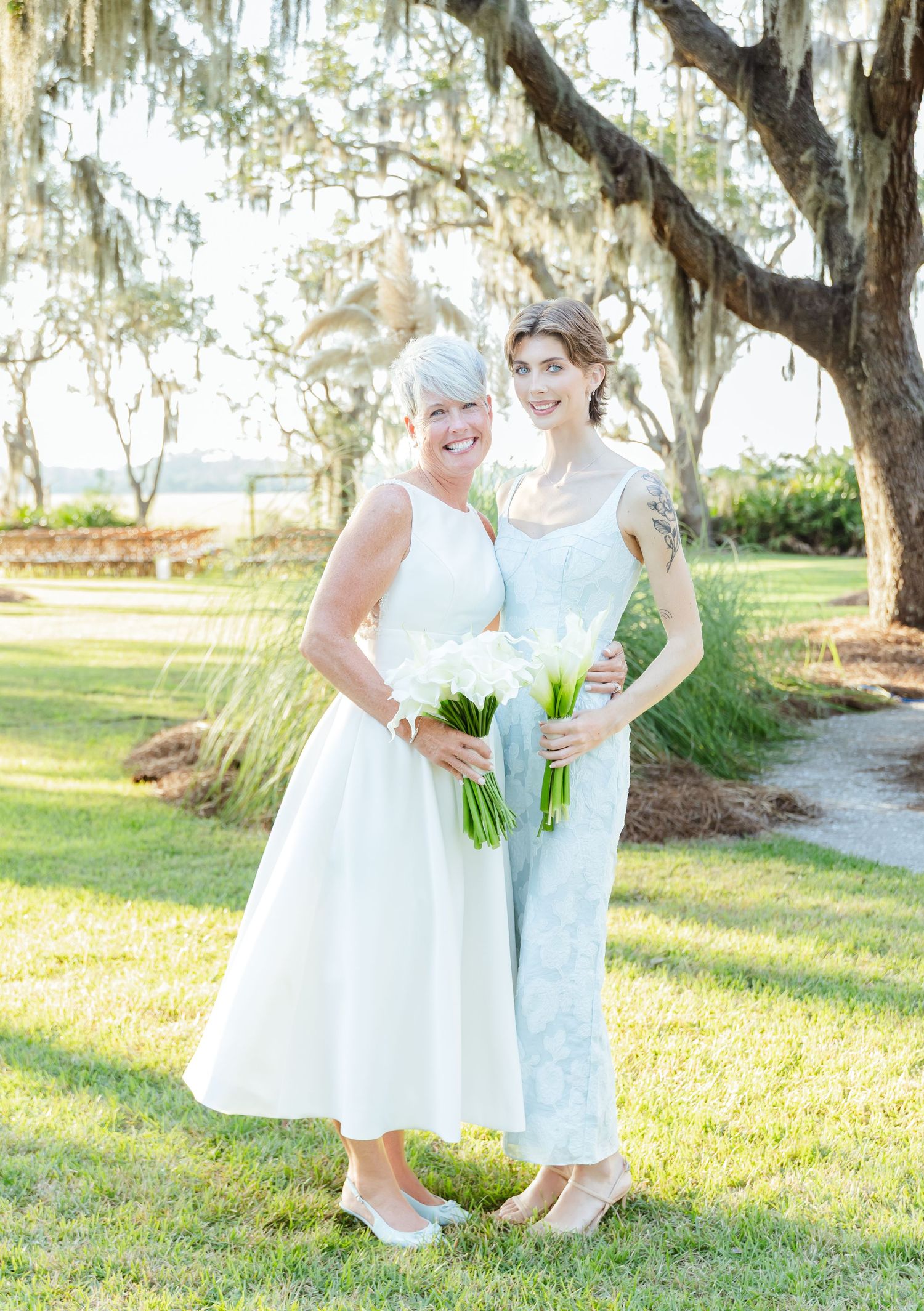 Two people in elegant dresses pose with white bouquets under Spanish moss draped trees at an outdoor venue.