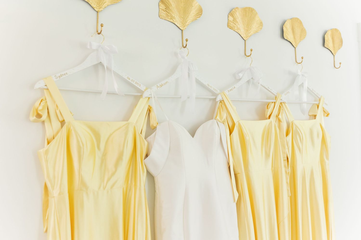 Pale yellow bridesmaid dresses hanging on white hangers against a white wall with gold leaf decorations.
