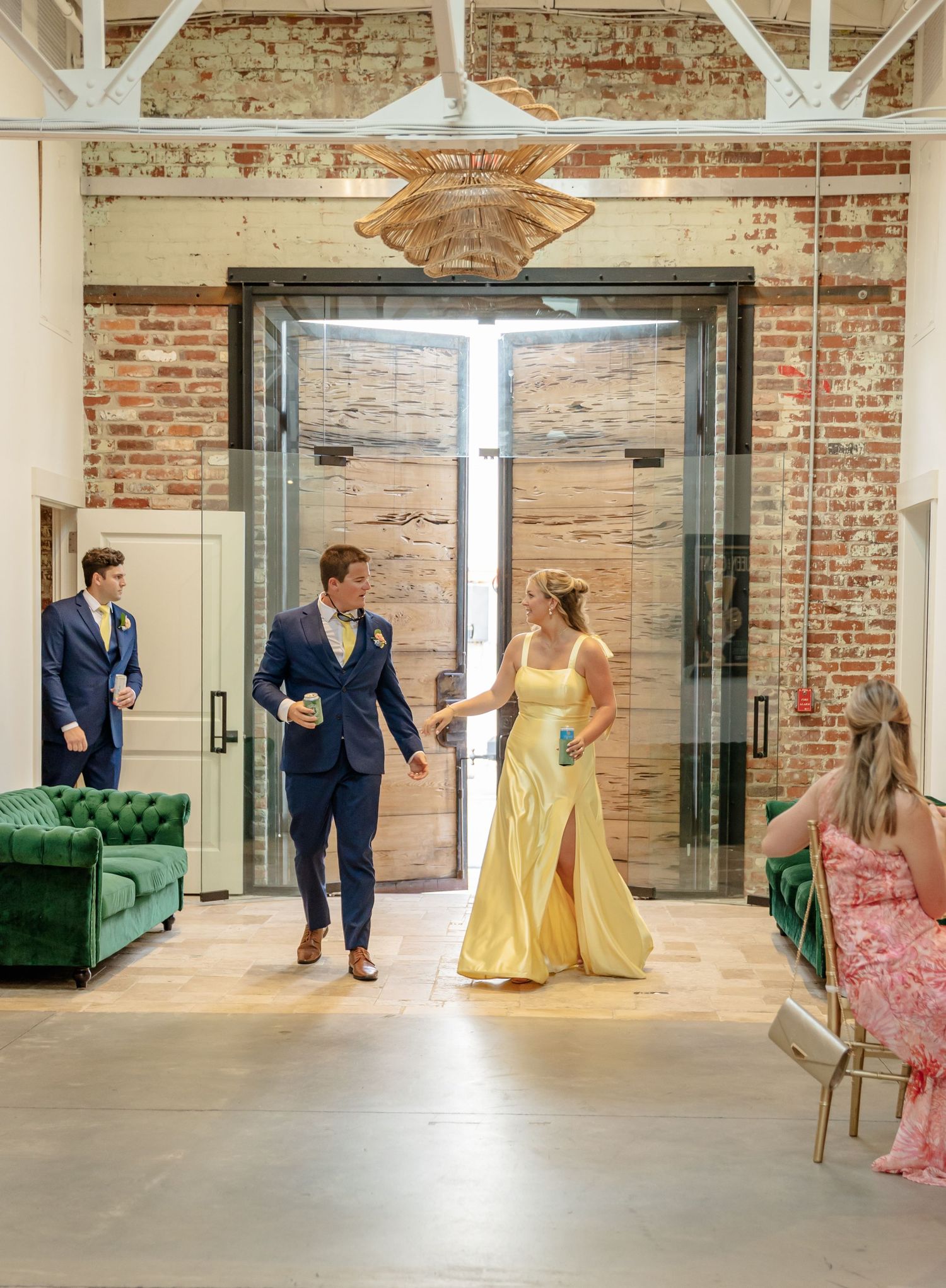 Wedding party in navy suits and yellow dresses dance in a rustic brick-walled venue with modern decor.