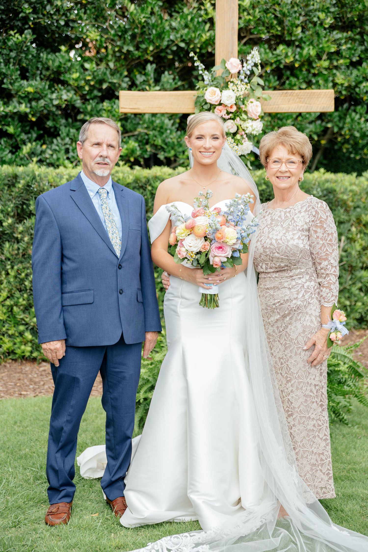 A wedding party poses outdoors in a garden setting with blue suits and white gowns against lush greenery.