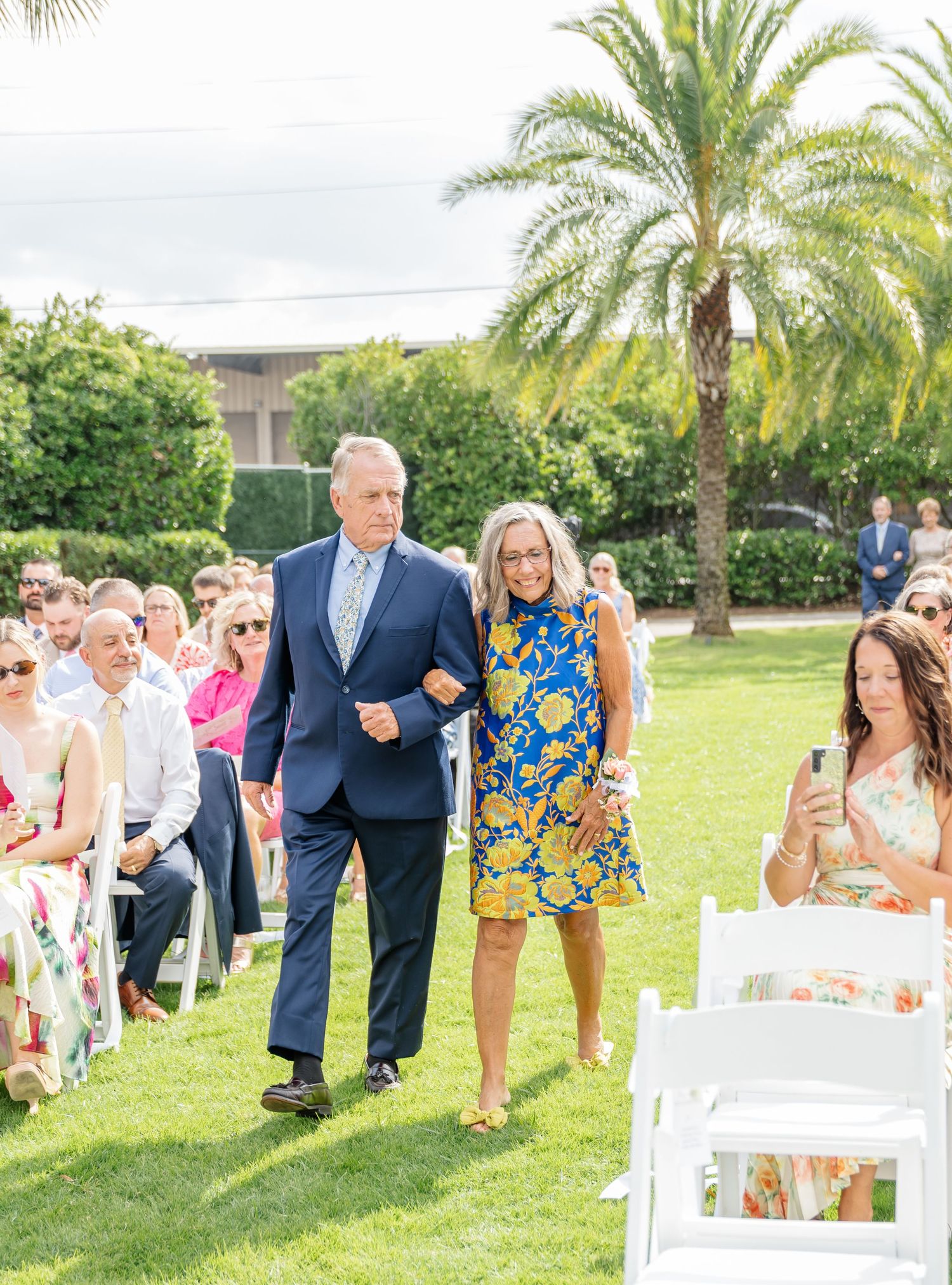 Wedding guests walking down an aisle lined with white chairs on a sunny lawn with palm trees in the background.