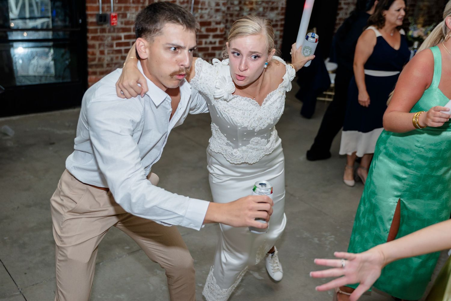 Wedding guests perform dance moves with enthusiasm at a rustic brick venue reception.