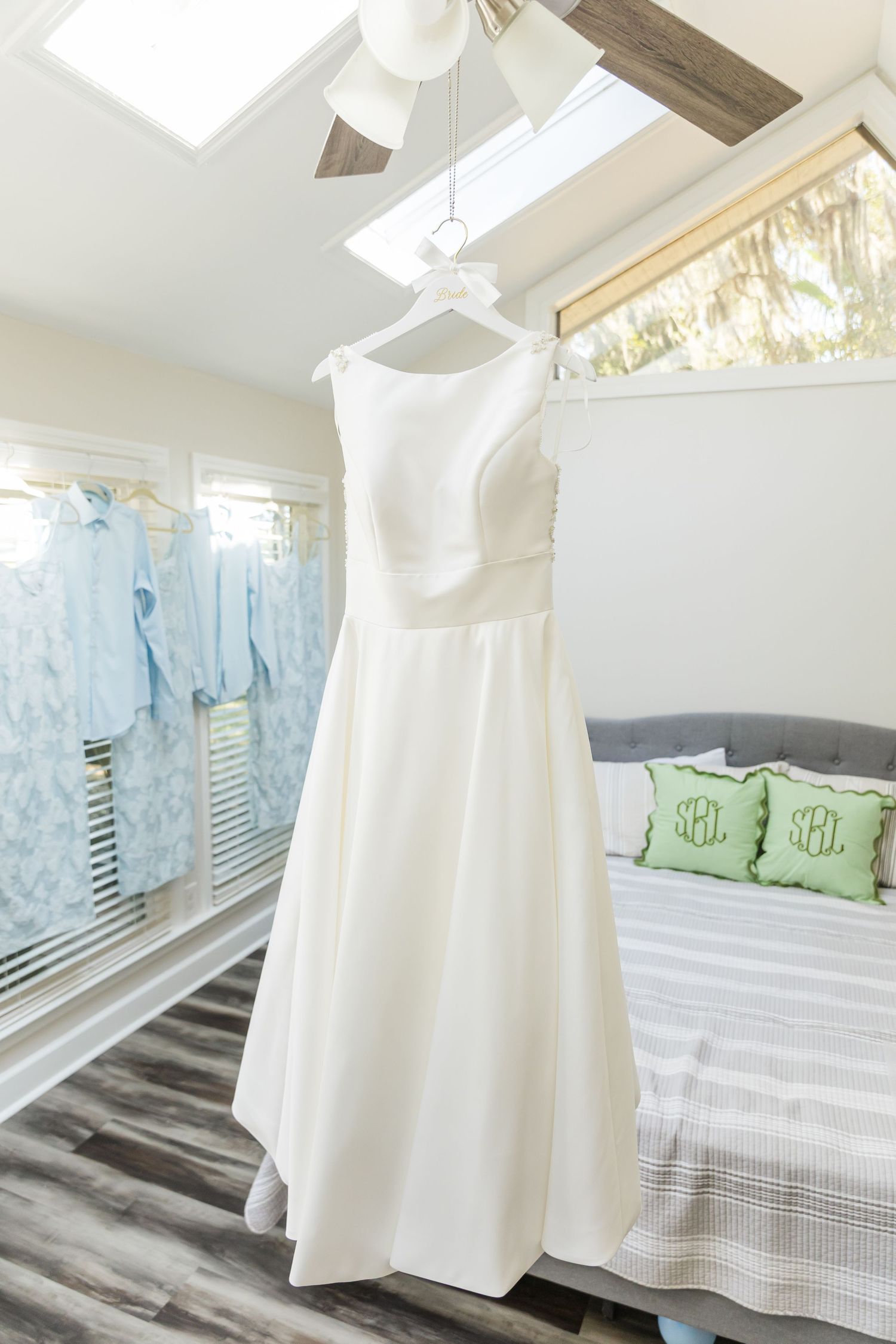 White baby dresses hanging on wooden hangers in a bright nursery with light blue curtains and green pillows.
