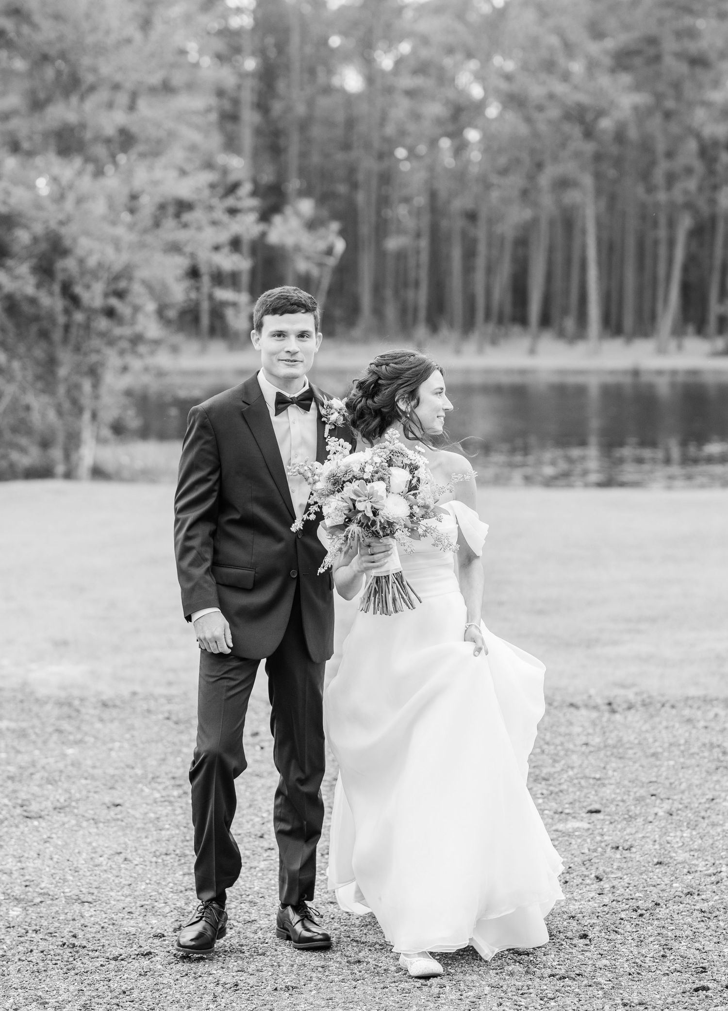 Black and white photo of a couple in wedding attire running playfully across a grassy field near trees.