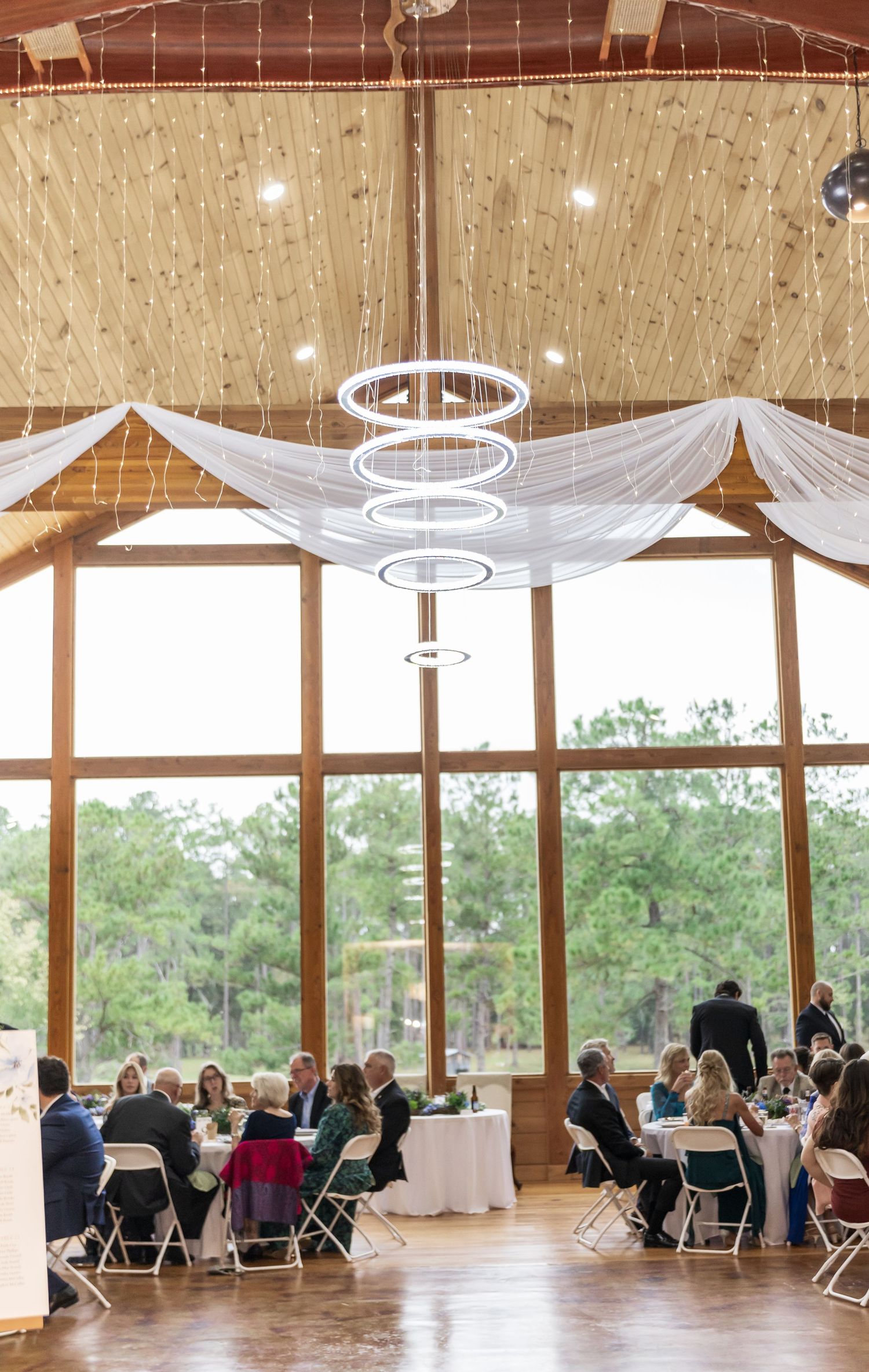 Modern rustic wedding reception venue with white draping and circular chandeliers overlooking a scenic forest view.