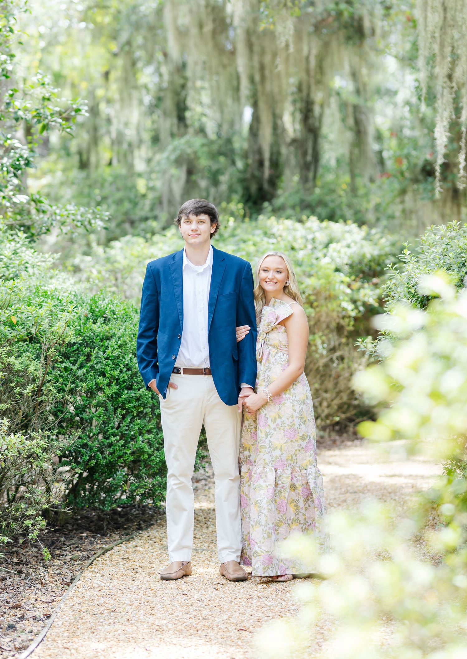 A couple walks together on a garden path surrounded by Spanish moss and lush greenery on a bright sunny day.