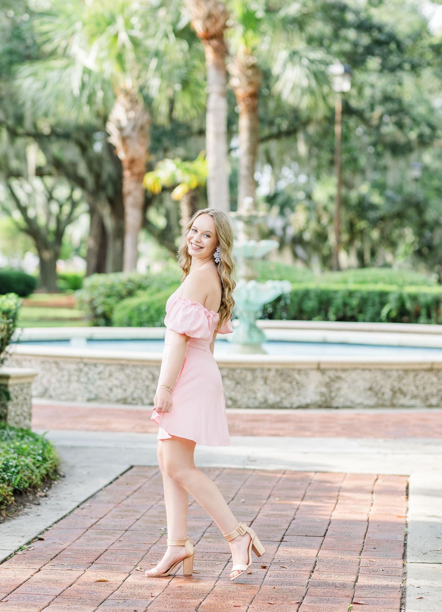 A soft pink dress flows gracefully against a backdrop of palm trees and manicured gardens.