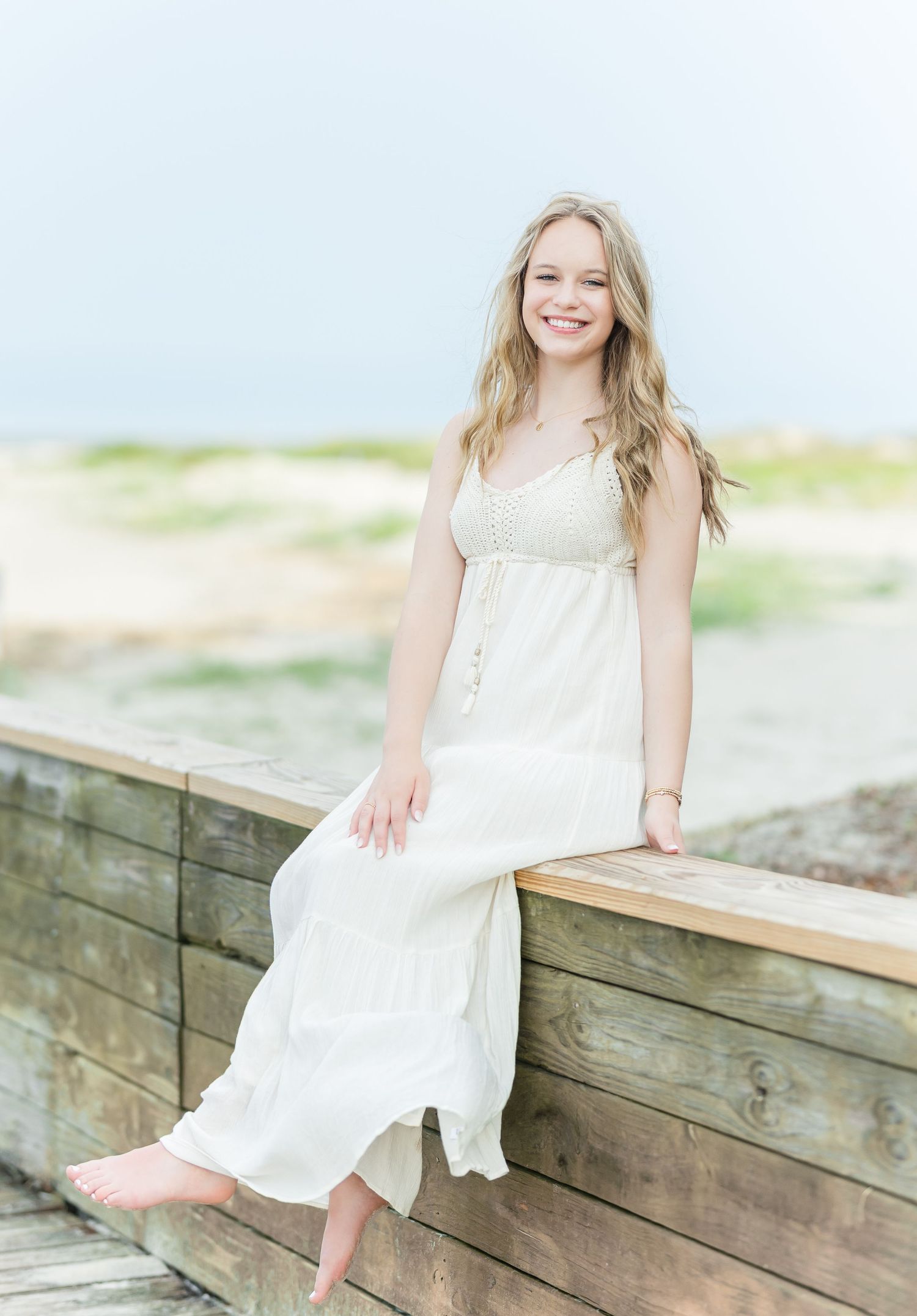 Person in white dress sitting on wooden boardwalk near beach during bright sunny day.
