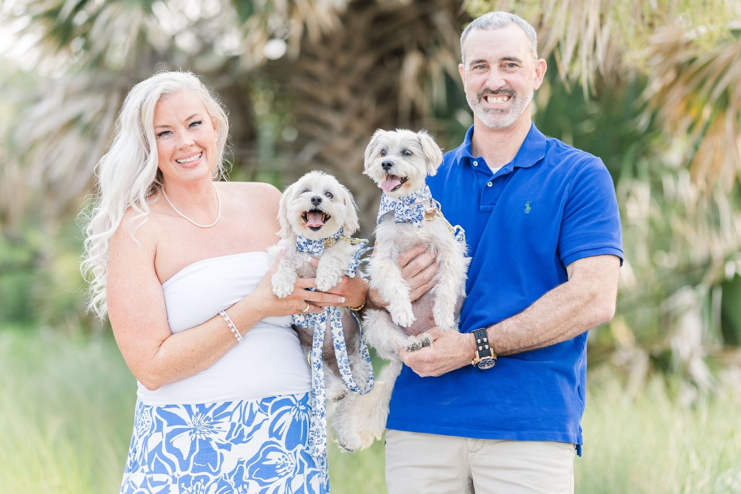 A couple in coordinating blue and white outfits holding their two small white dogs on a beach.