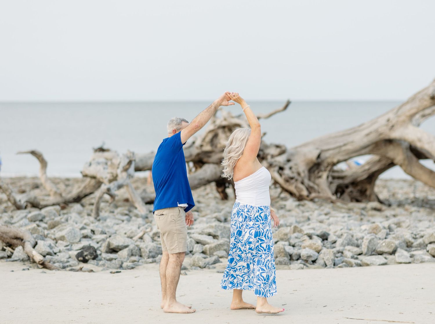 A couple dances together on a beach with driftwood and the ocean in the background.