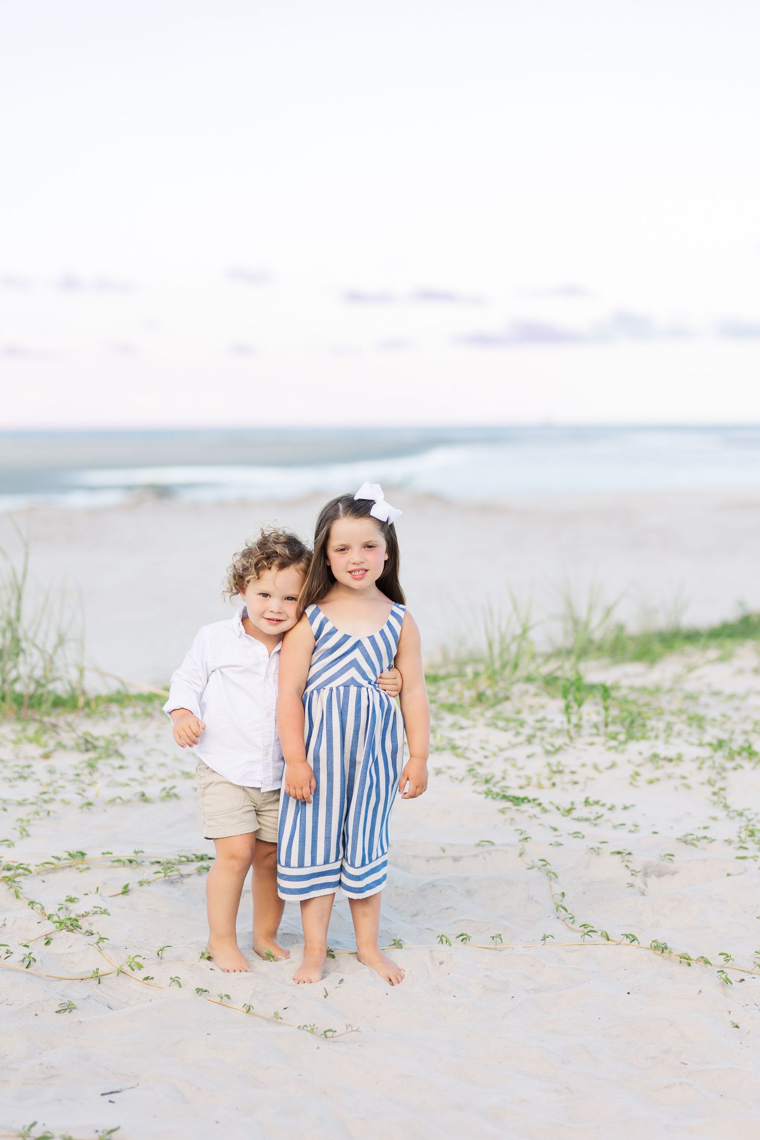 Two small children standing together on a sandy beach with ocean waves in the background.