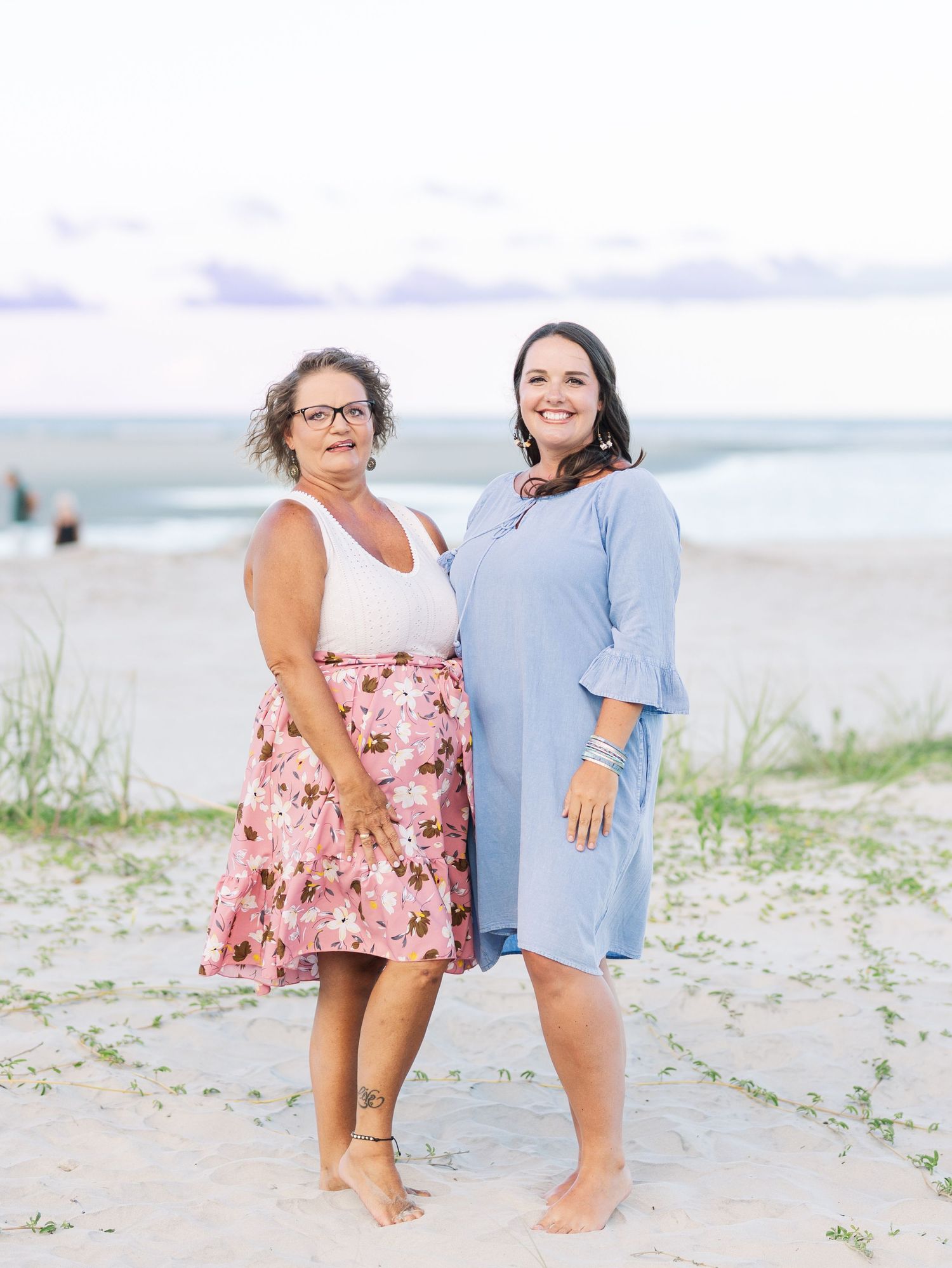 Three people in summery outfits stand barefoot on a sunny beach with scattered beach grass.