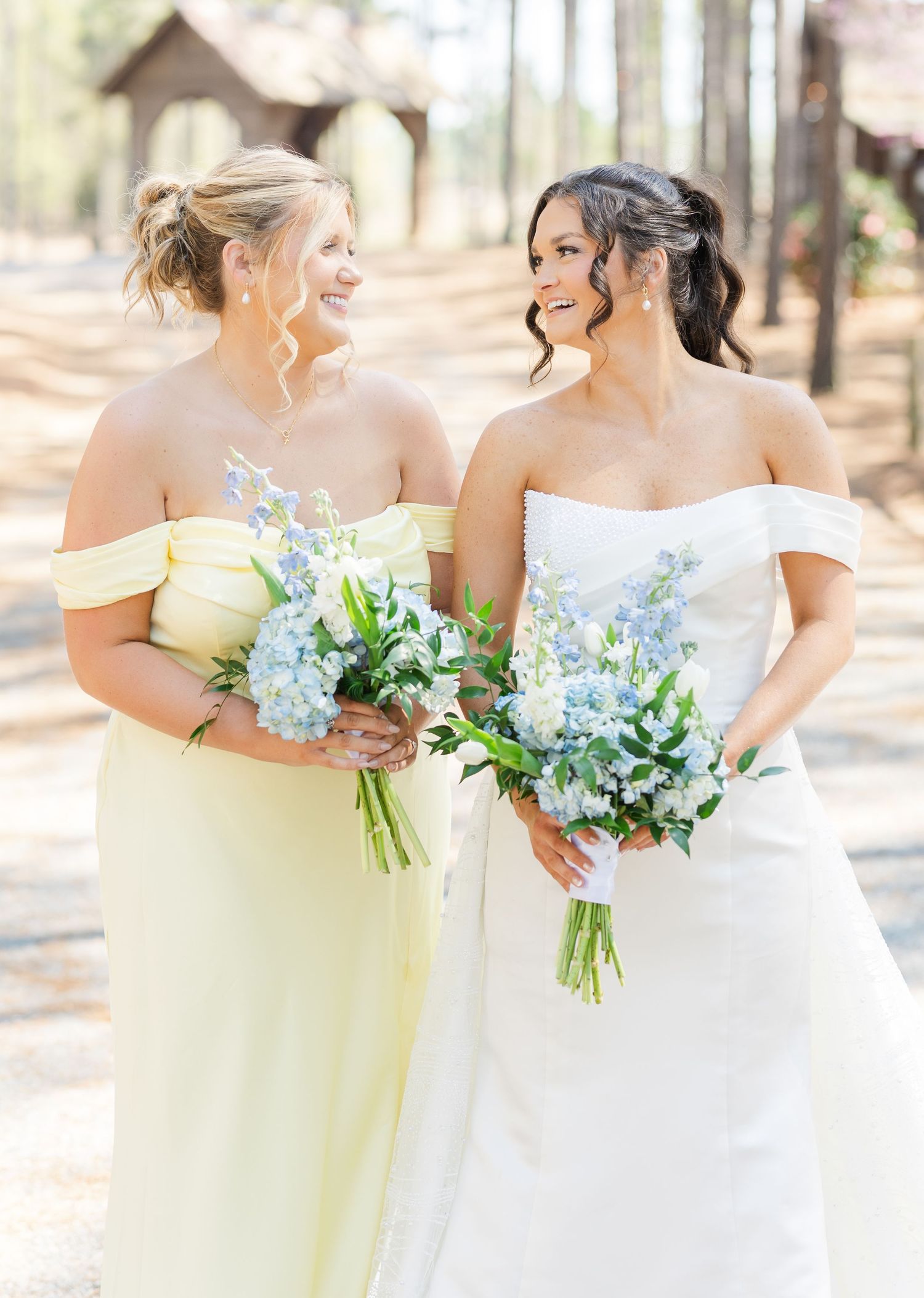 Bride in strapless white gown and bridesmaid in yellow off-shoulder dress hold blue bouquets outdoors.
