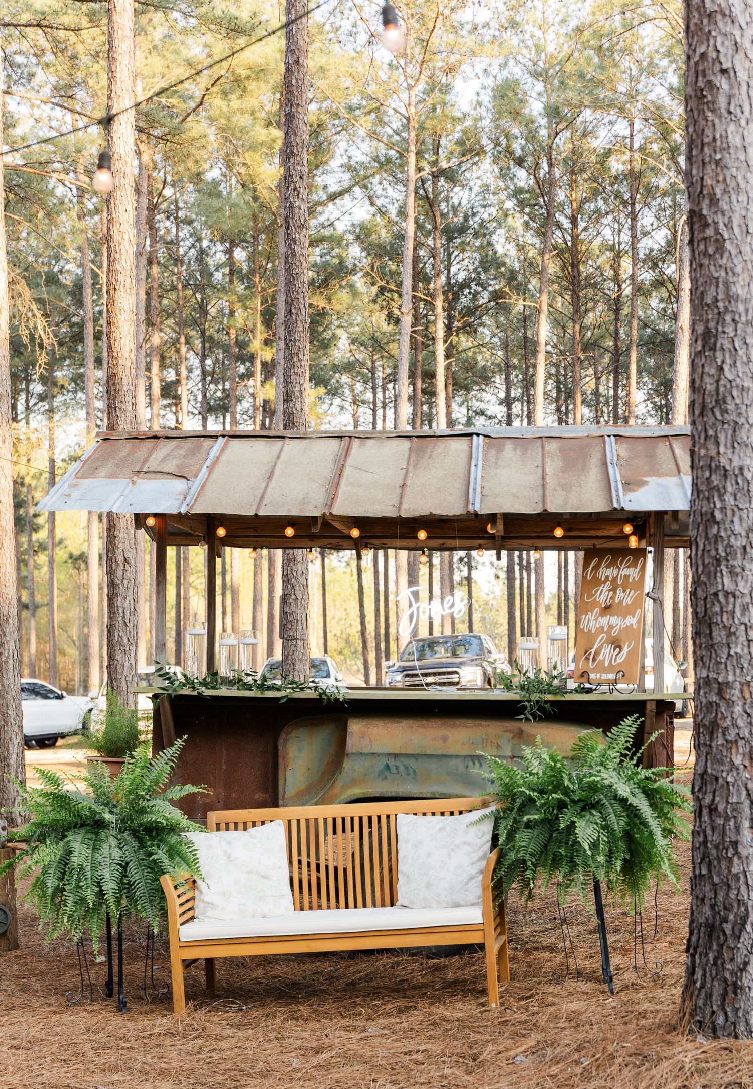Rustic outdoor wedding bar with tin roof, string lights, ferns, and a wooden bench in pine forest.