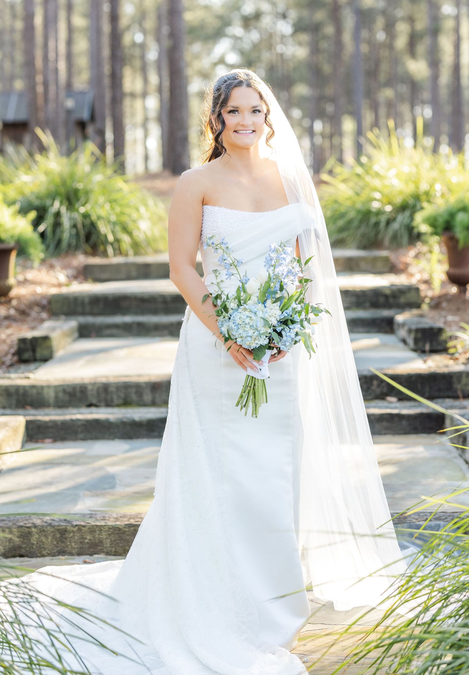 Elegant bride in strapless white gown holds blue hydrangea bouquet on stone steps outdoors.