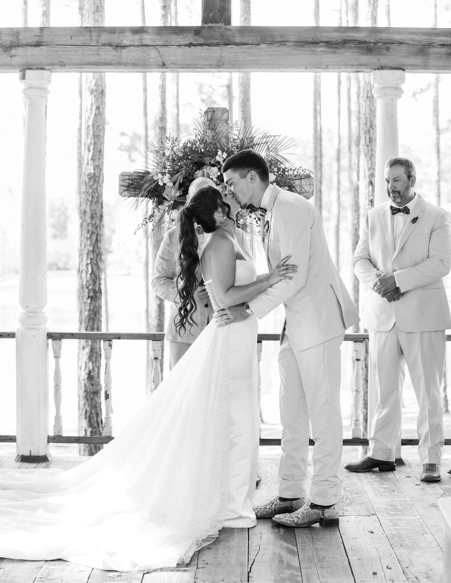 Bride and groom share first kiss at rustic outdoor altar as officiant watches in black and white photo.