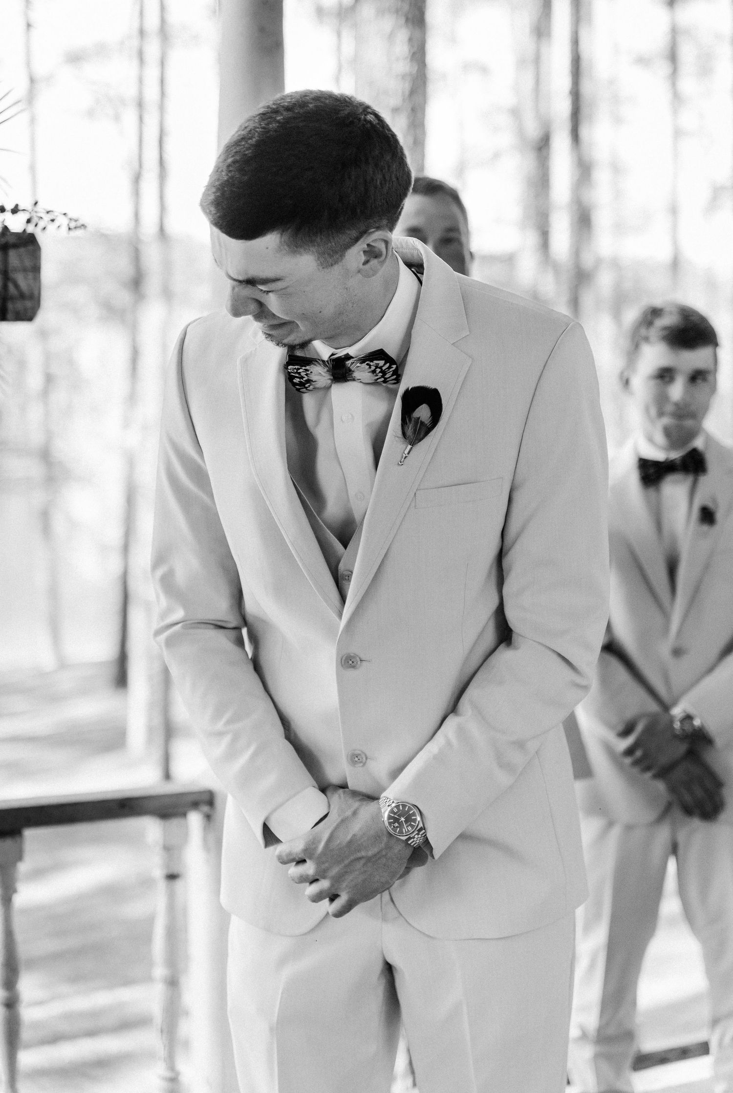 Emotional groom in white suit with floral bow tie clasping hands at wedding altar in black and white photo.
