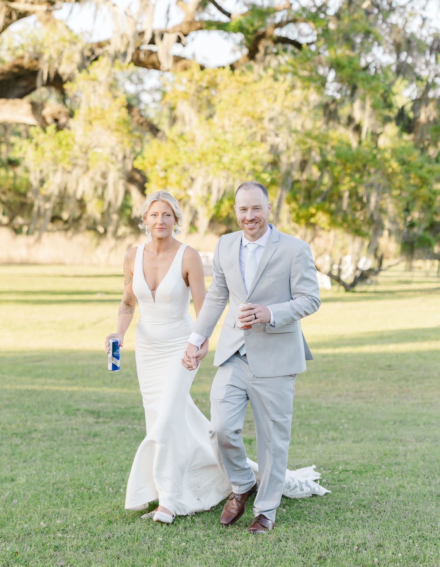 Smiling bride and groom walking hand in hand on green lawn under Spanish moss trees.
