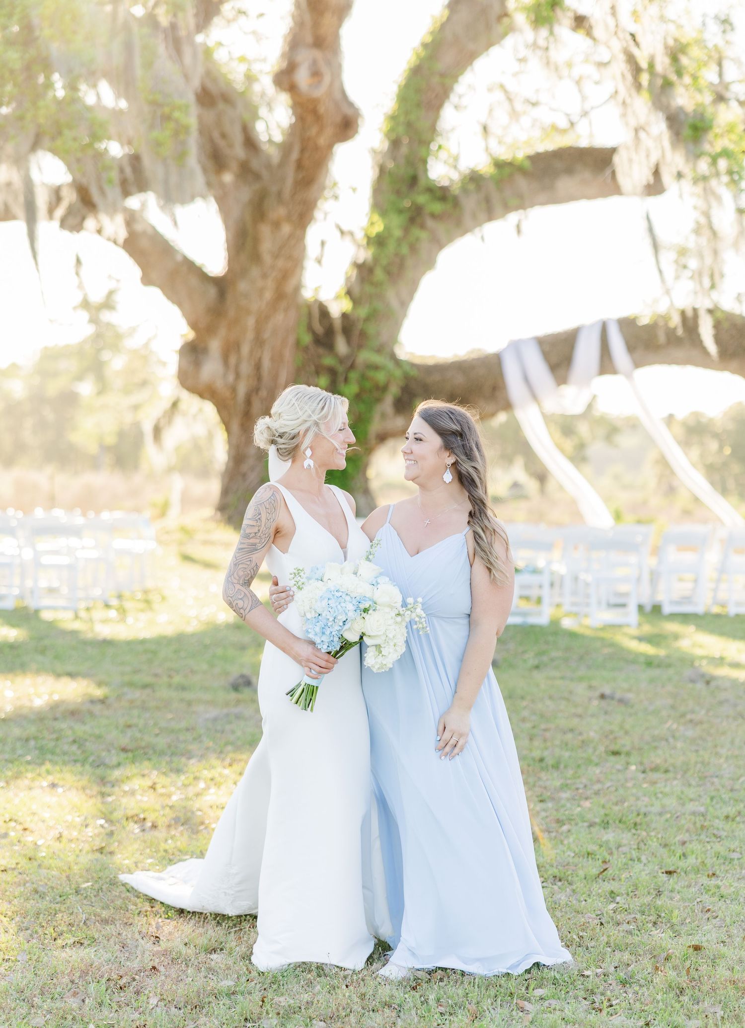 Bride and bridesmaid in light blue dress sharing a smile under a moss-draped oak tree.