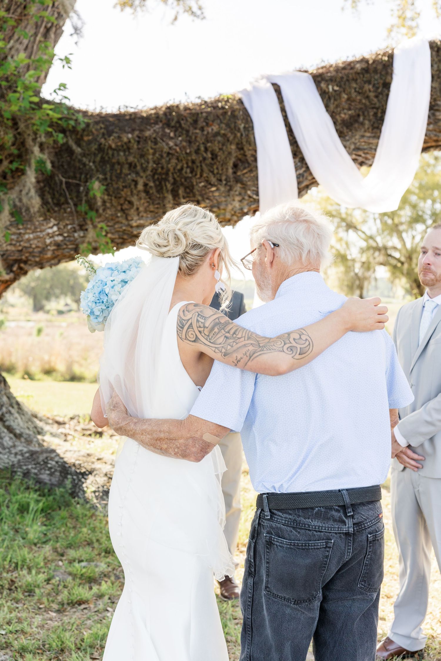 Bride with tattoo sleeve embraces elderly man at outdoor wedding ceremony under oak tree with white draping.