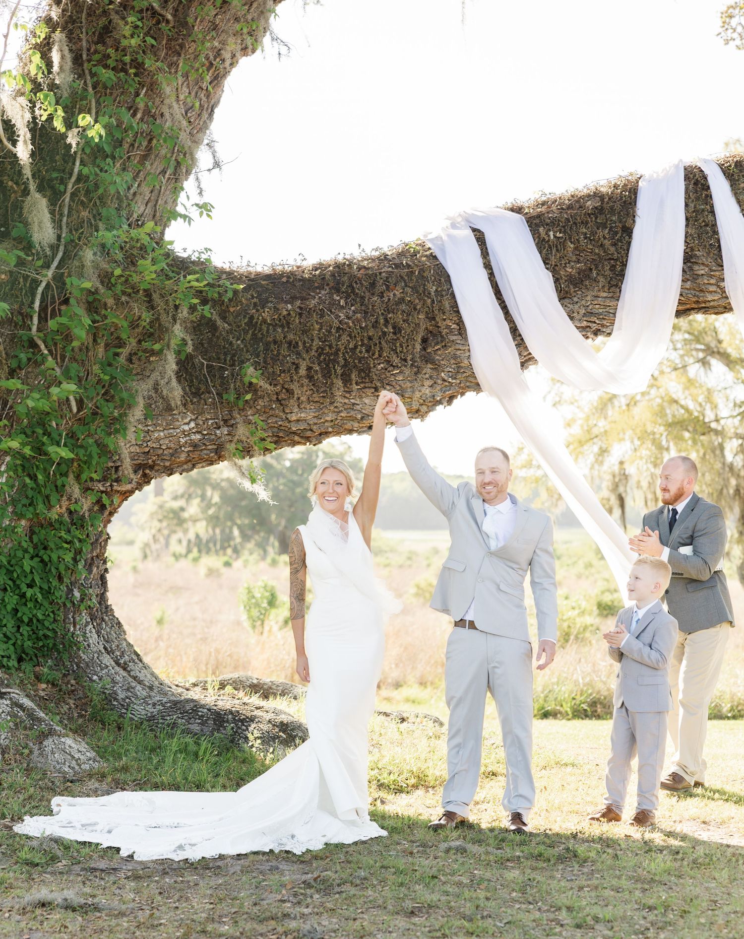 Newlywed couple raises hands in celebration under a moss-draped oak tree at outdoor wedding ceremony.